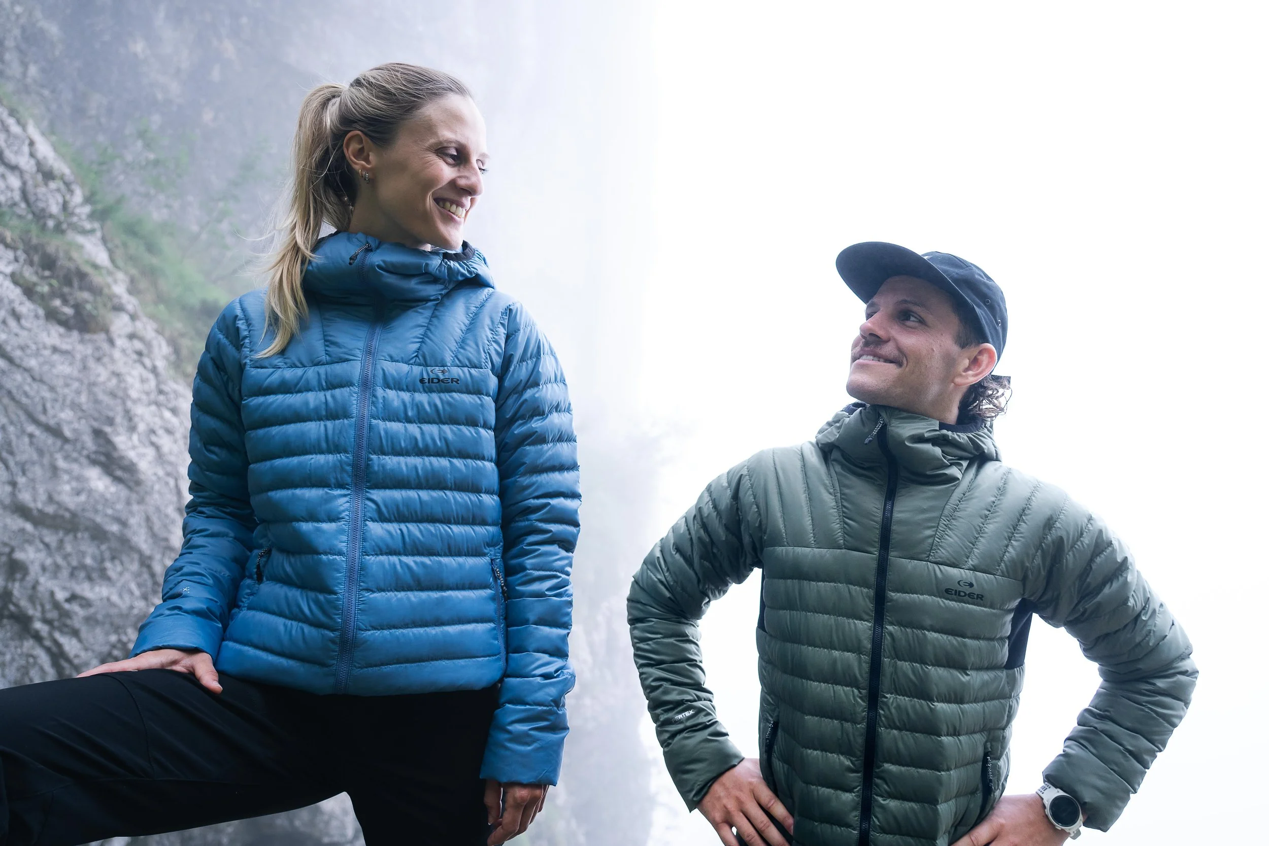 Deux personnes souriant en pleine nature, portant des vestes matelassées de couleurs différentes, une bleue et une verte, avec des montagnes en arrière-plan.