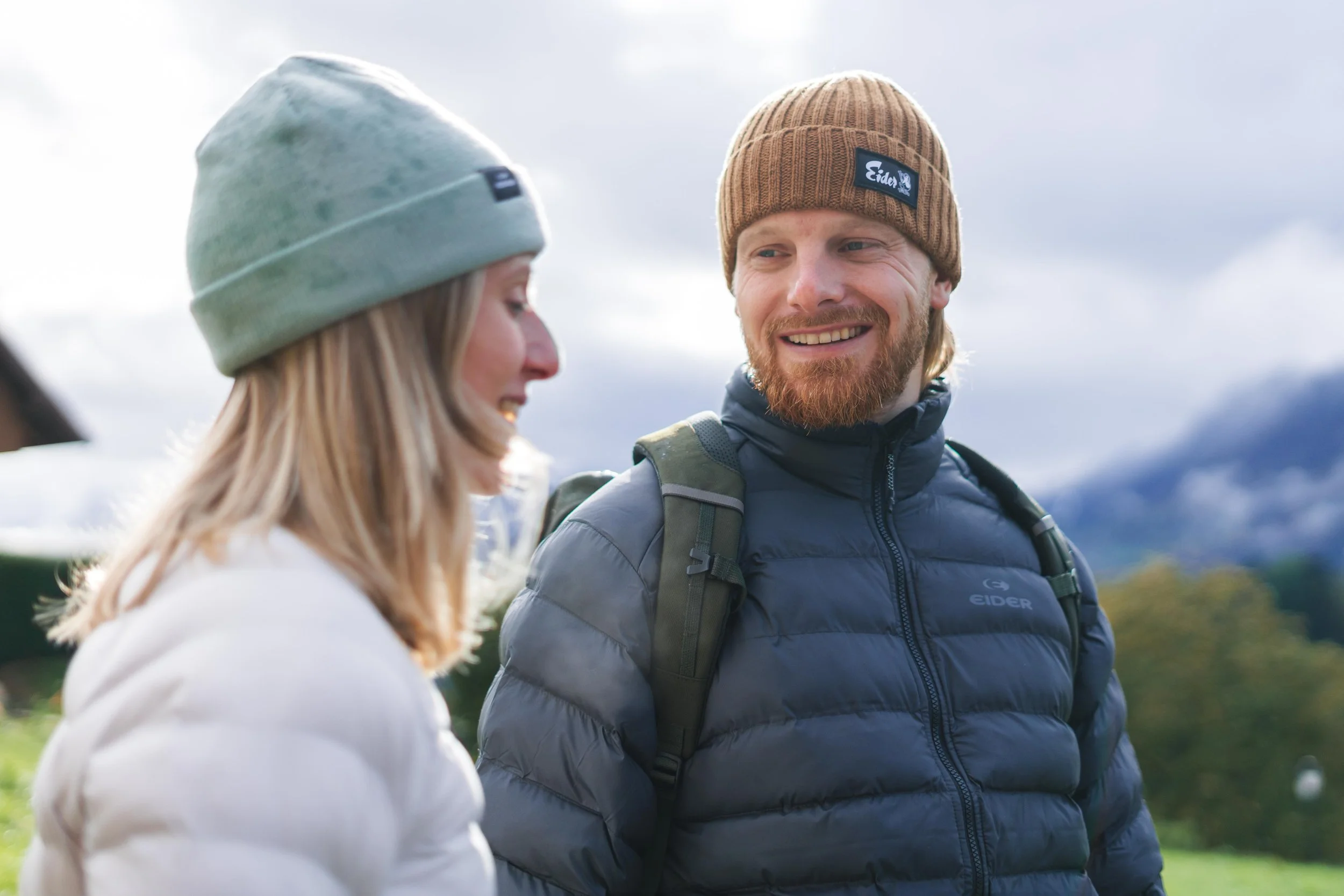 Deux personnes portant des bonnets et des manteaux d'hiver se sourient en plein air dans un paysage naturel.