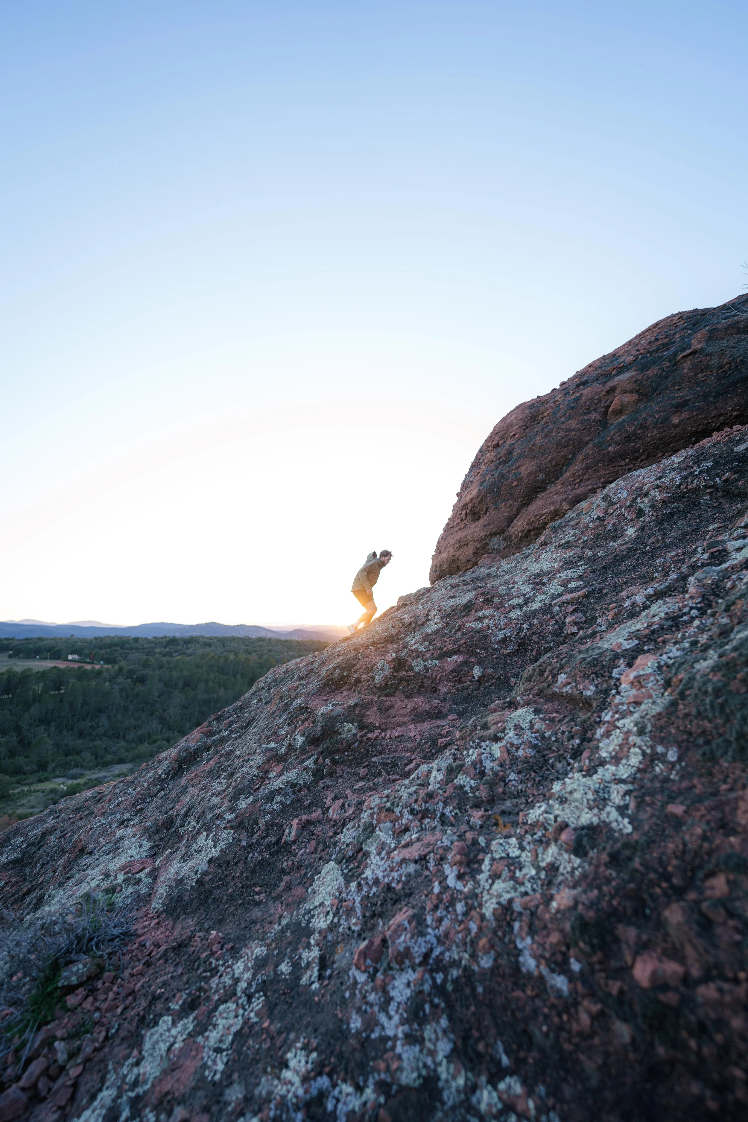 Personne escaladant une colline rocheuse au coucher du soleil avec une vue sur une forêt et des montagnes à l'arrière-plan.