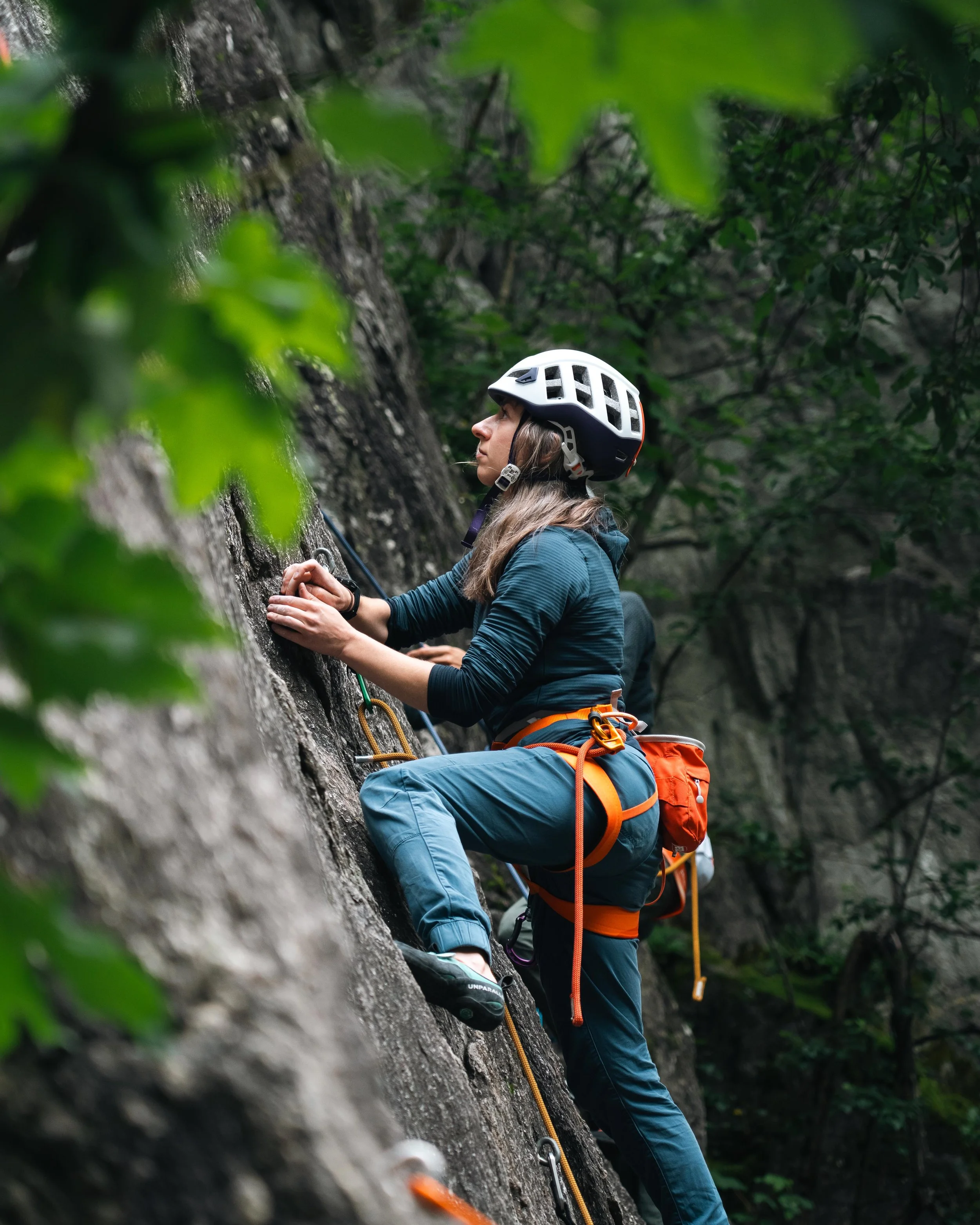Une personne escalade une paroi rocheuse avec un équipement de sécurité, portant un casque et un harnais orange, entourée de végétation.