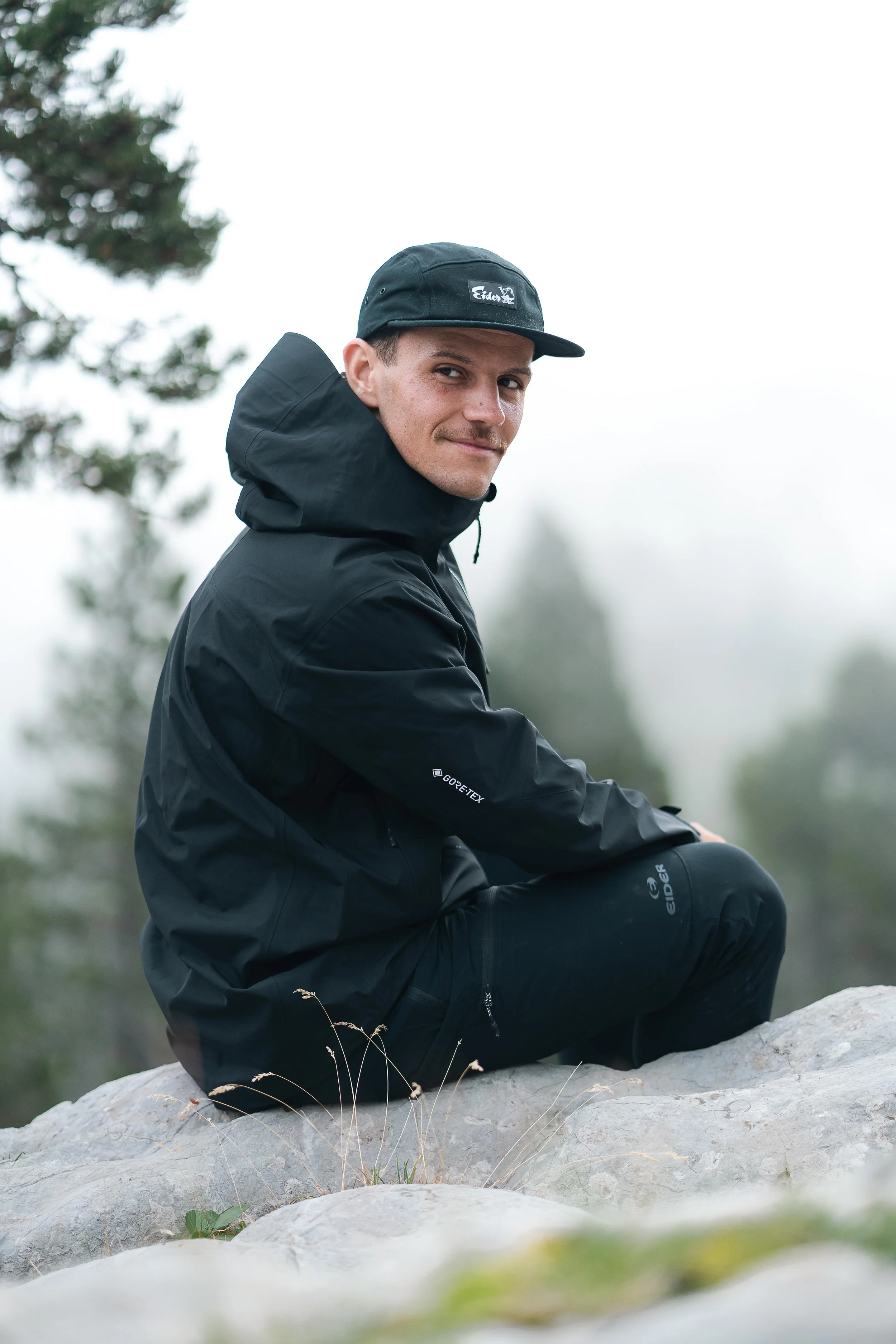 Un homme portant une veste et une casquette noires assis sur un rocher dans un environnement naturel flou.