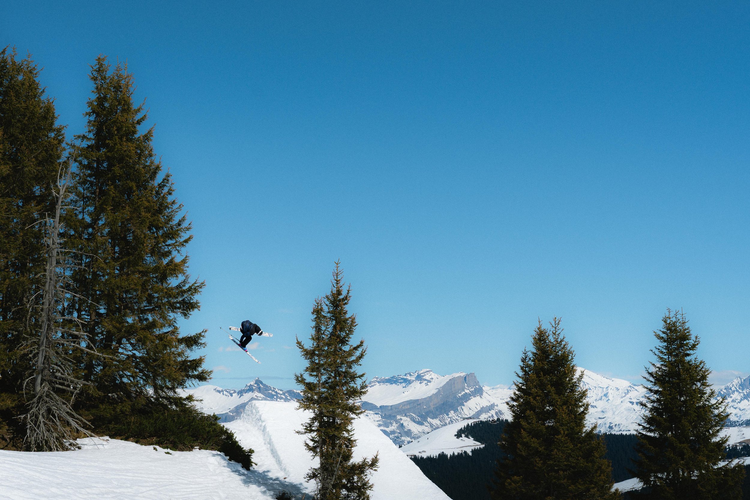 Skieur effectuant un saut dans une station de ski avec des montagnes enneigées et des arbres en arrière-plan, sous un ciel bleu.