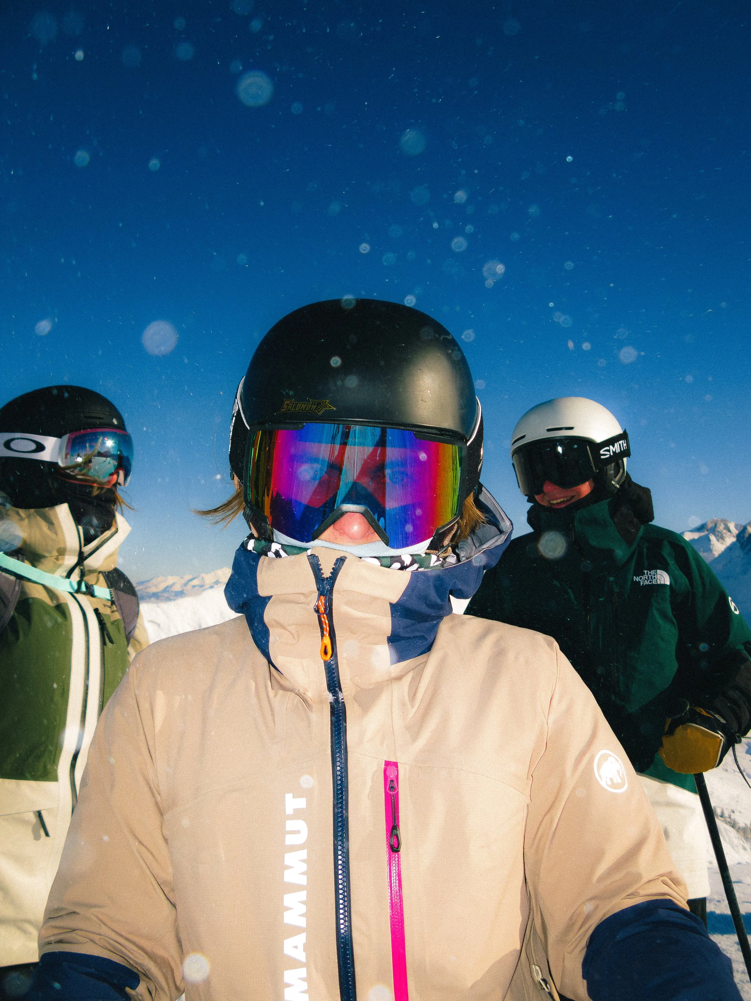 Trois personnes en tenue de ski sur une montagne enneigée sous un ciel bleu. Elles portent des casques, des lunettes et des vestes de ski.