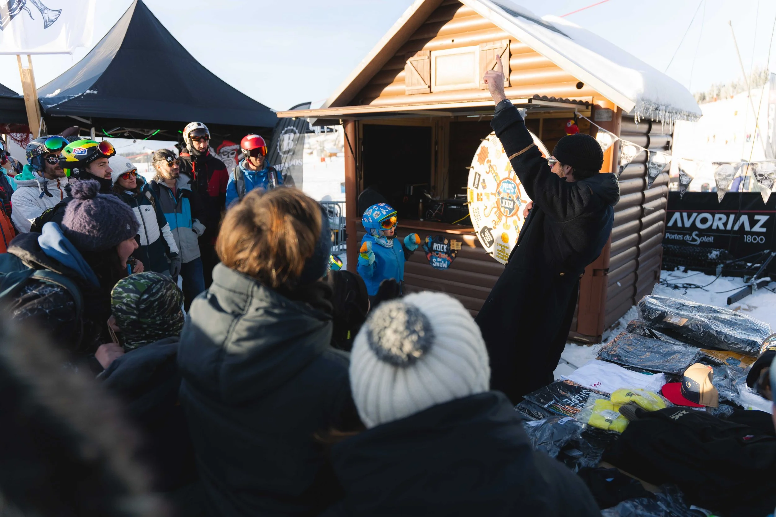 Un groupe de personnes habillées pour le ski ou la neige se tient devant une petite cabane en bois. Un homme lève le bras vers une roue de la fortune, tandis qu'un enfant portant une combinaison bleue semble enthousiaste. Des tentes et des produits s