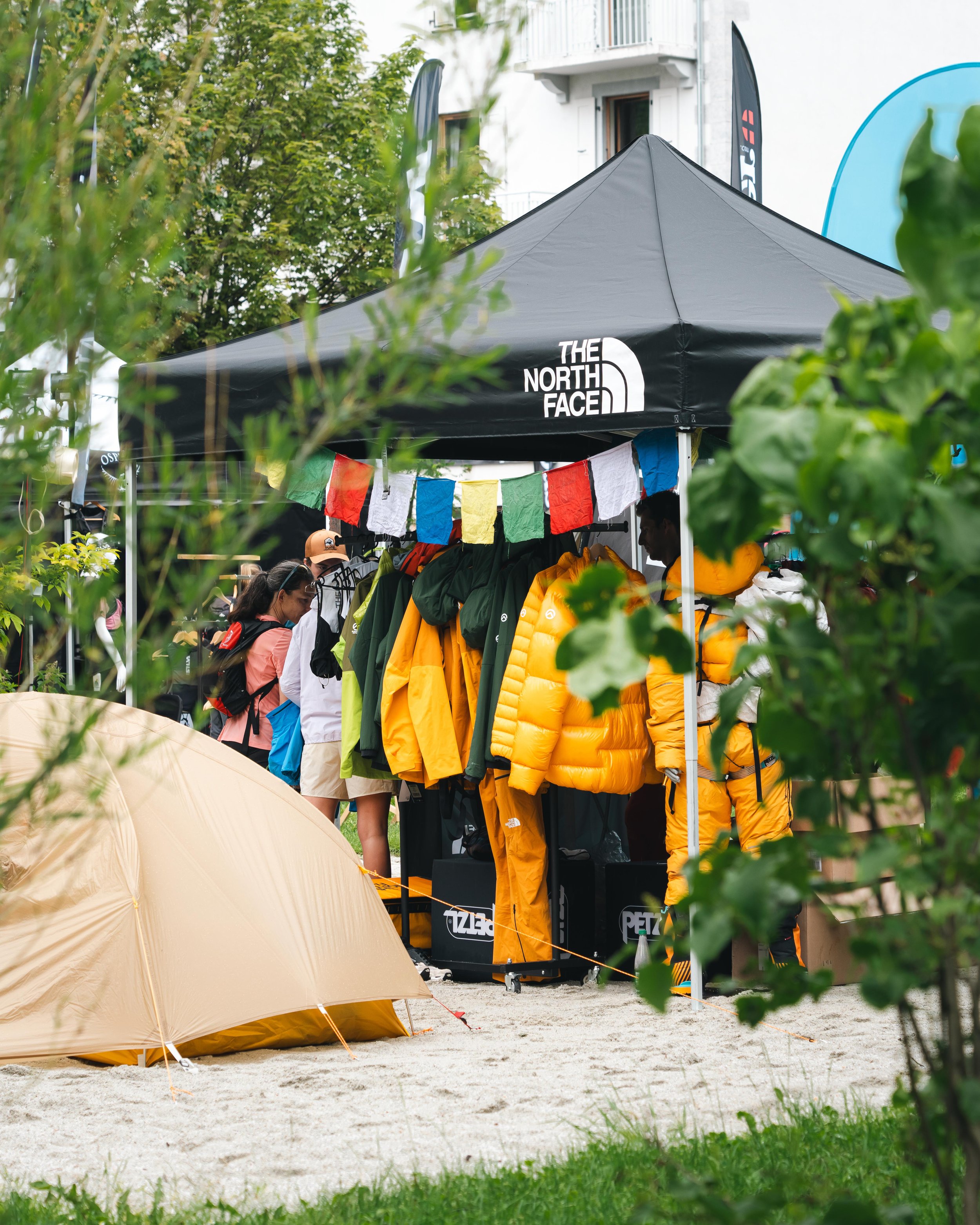 Un stand de The North Face dans une foire extérieure, avec une tente beige et des vêtements d'extérieur colorés en exposition. Des personnes sont présentes autour du stand, entourées de verdure.