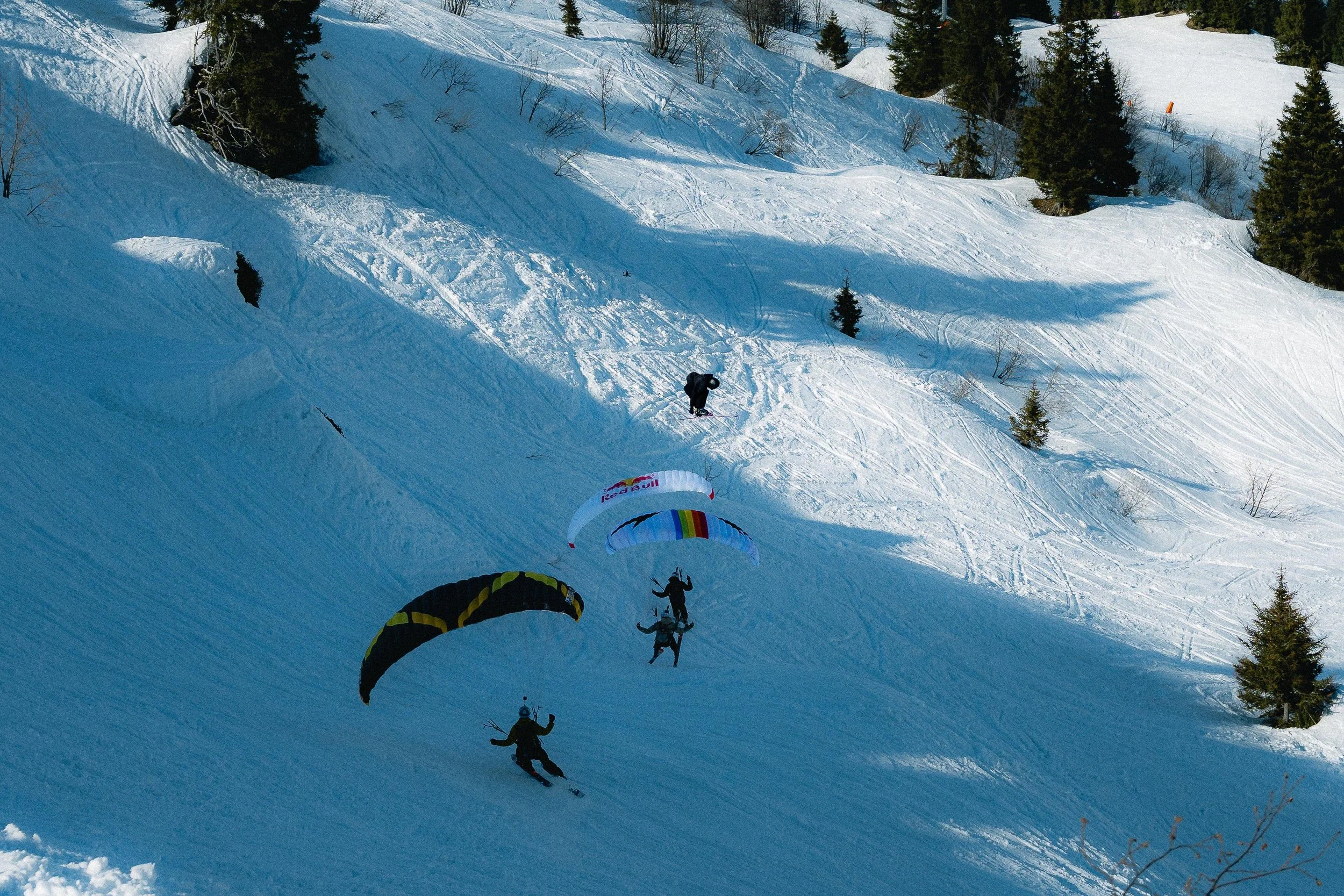 Skieurs en speedriding sur une pente enneigée avec des voiles colorées, entourés de quelques arbres et un ciel clair.