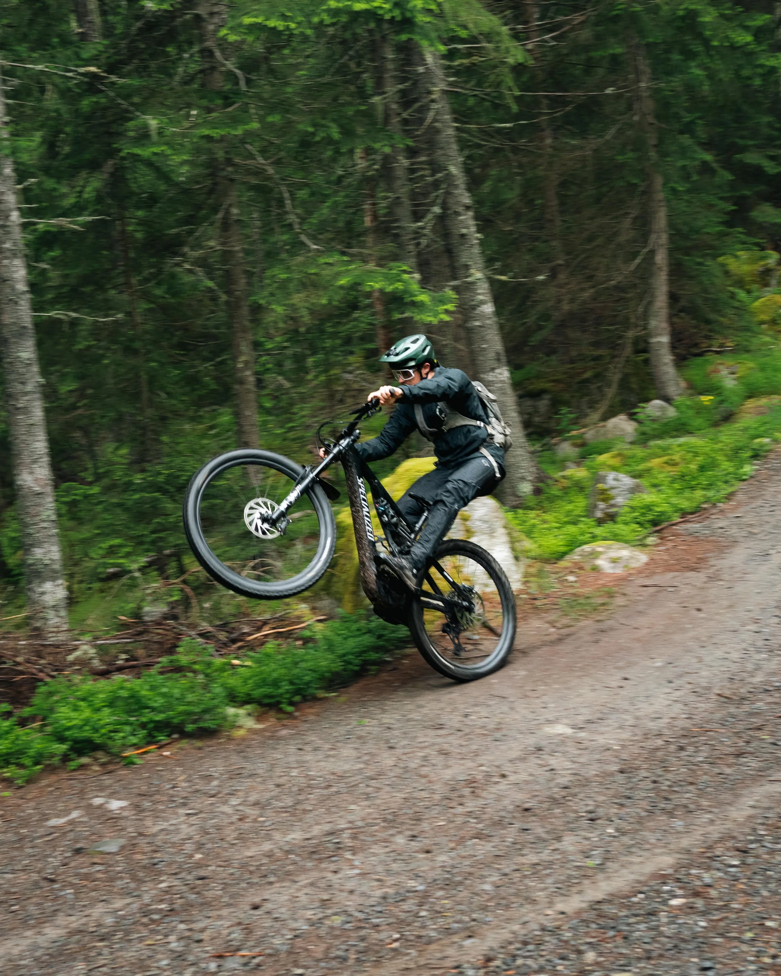 Personne sur un vélo tout-terrain dans une forêt, faisant un wheeling sur un sentier en gravier.