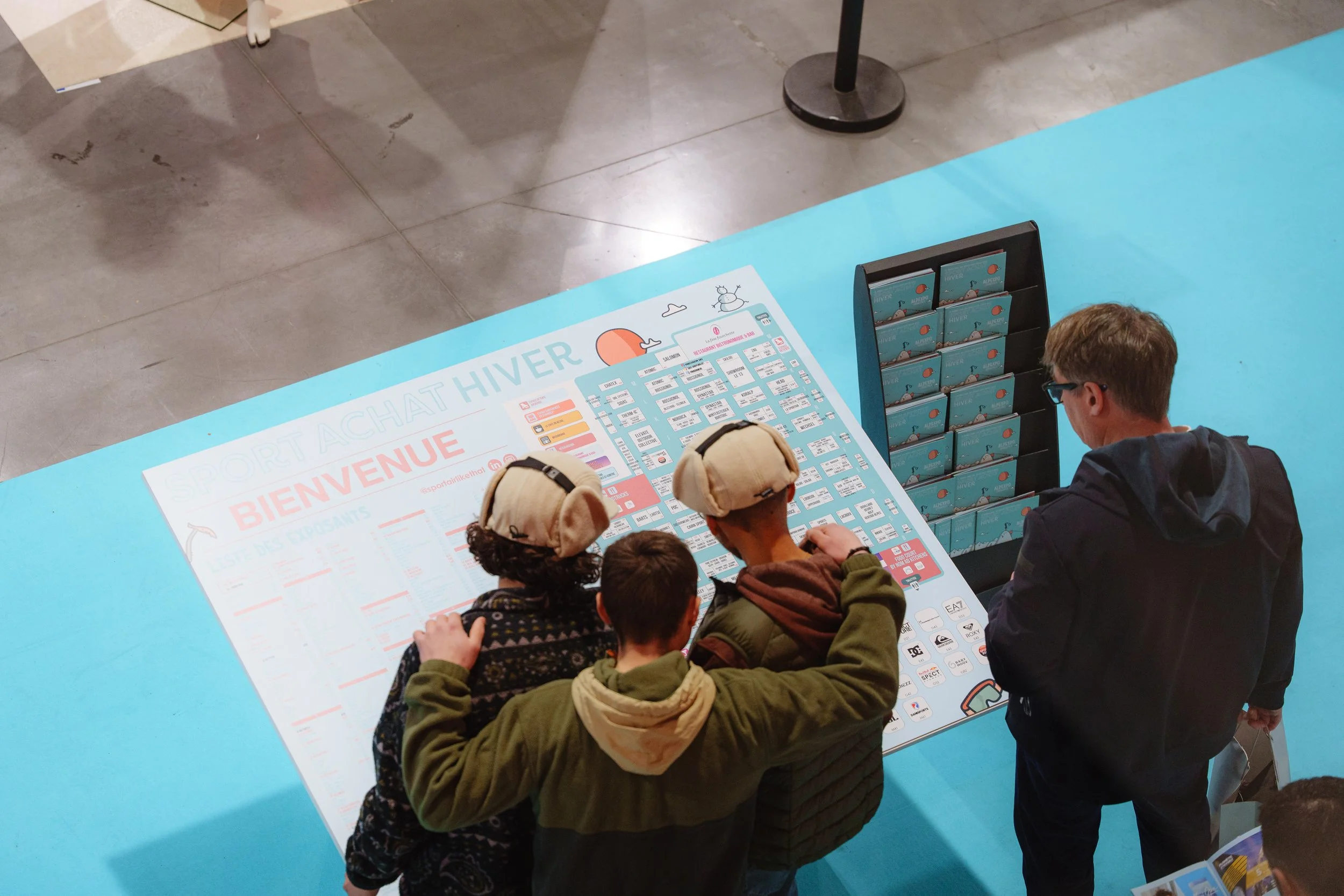 Des personnes regardent un plan au sol d'une exposition intitulée "Sport Achat Hiver" avec des stands et une liste d'exposants.
