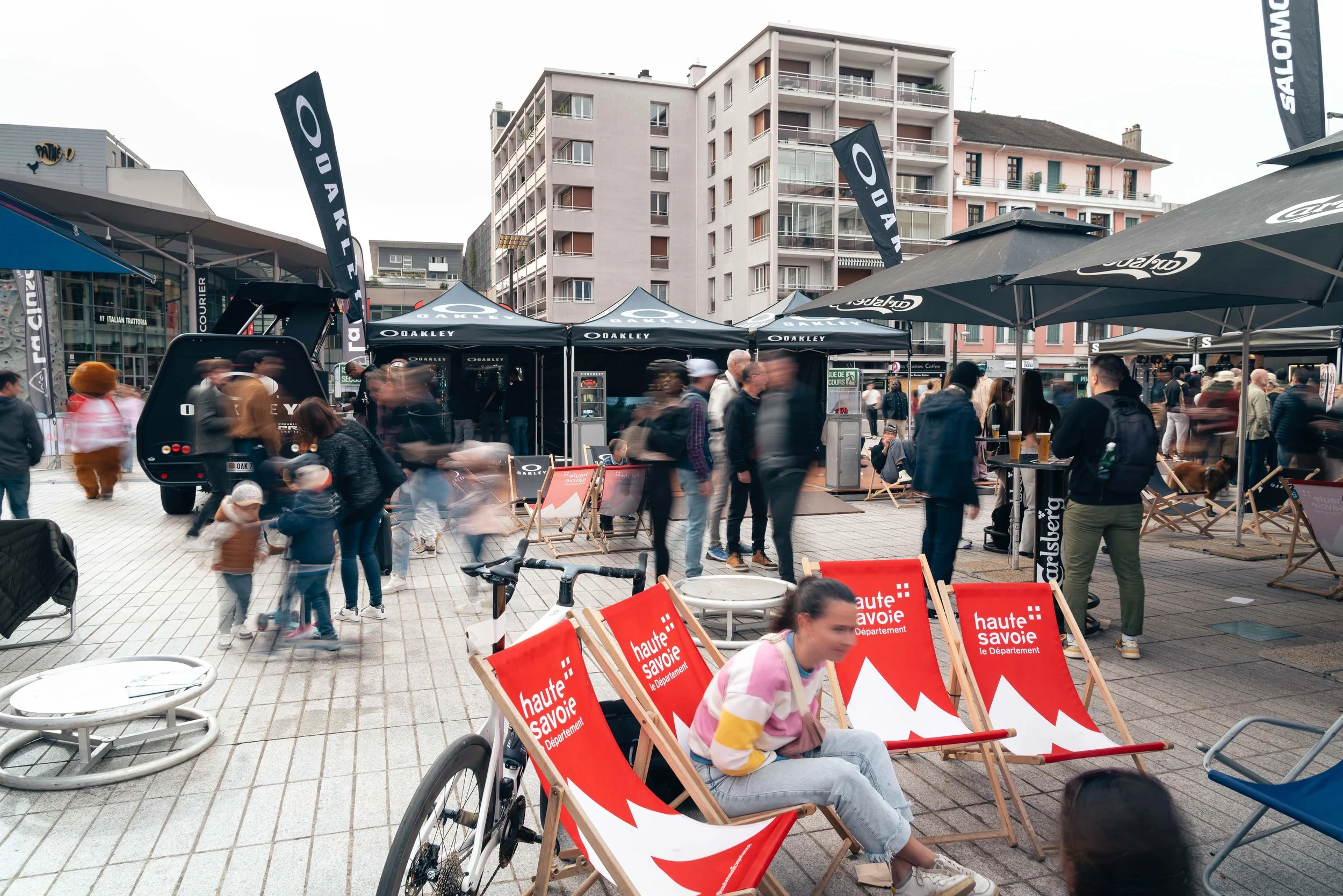 Un événement urbain avec des stands Oakley, des chaises rouges à l'effigie de la Haute-Savoie, des gens marchant et discutant, et des bâtiments en arrière-plan.