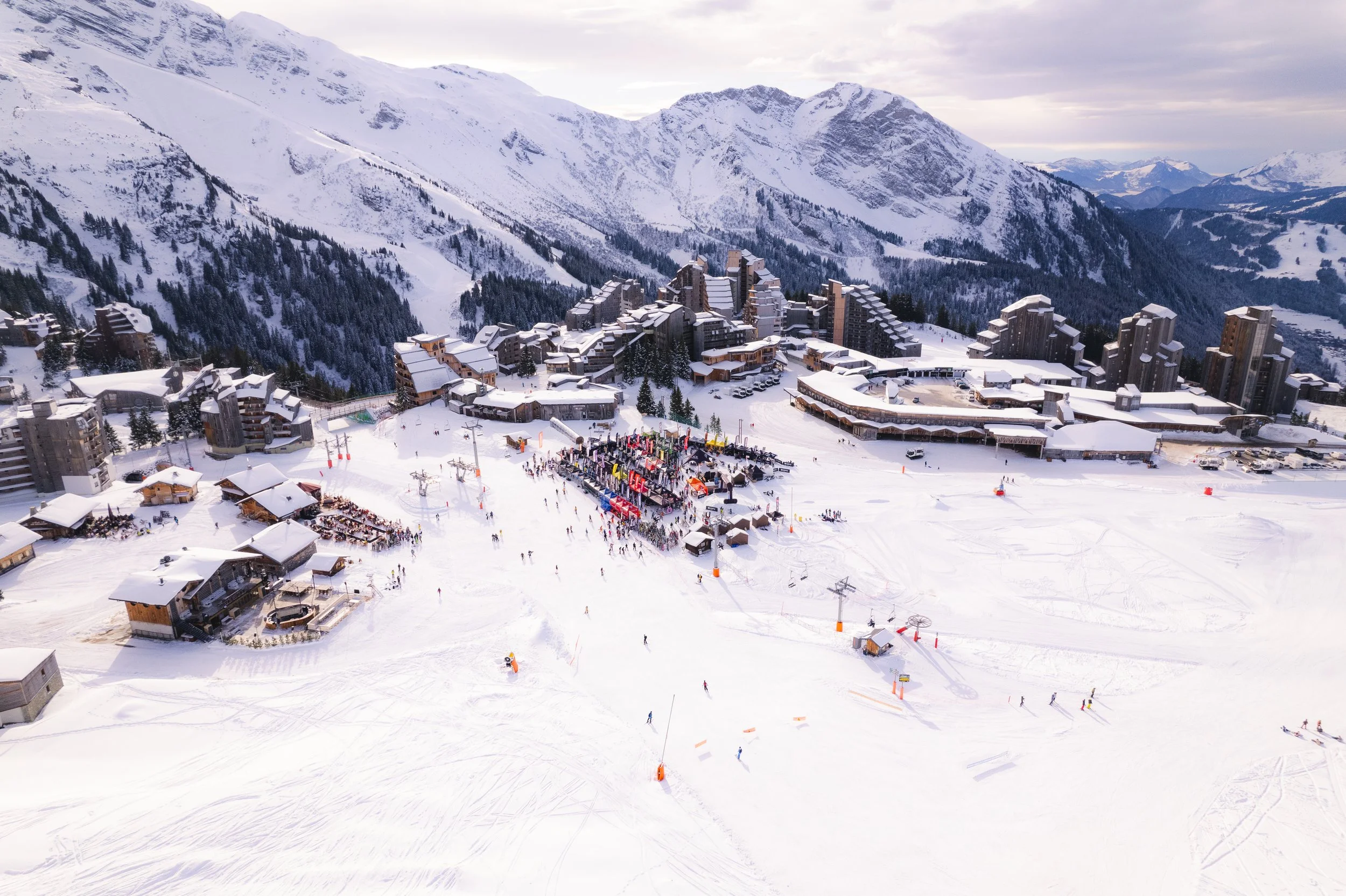 Vue aérienne d'une station de ski avec des montagnes enneigées, des bâtiments et des skieurs. Le paysage montre des pistes de ski animées et de la neige fraîche.