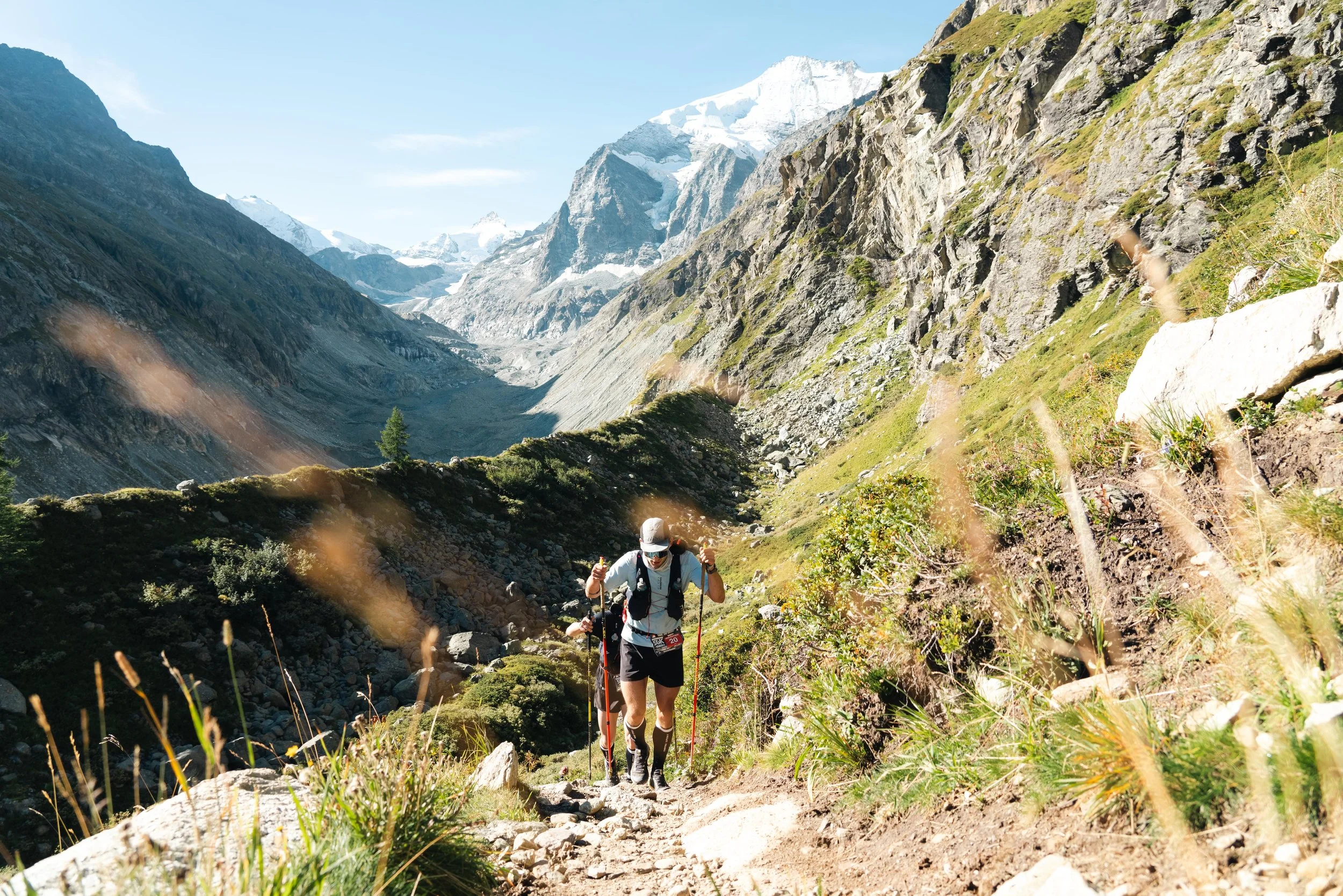 Deux randonneurs gravissent un sentier montagneux rocheux avec des bâtons de marche, entourés de montagnes et de végétation alpine.