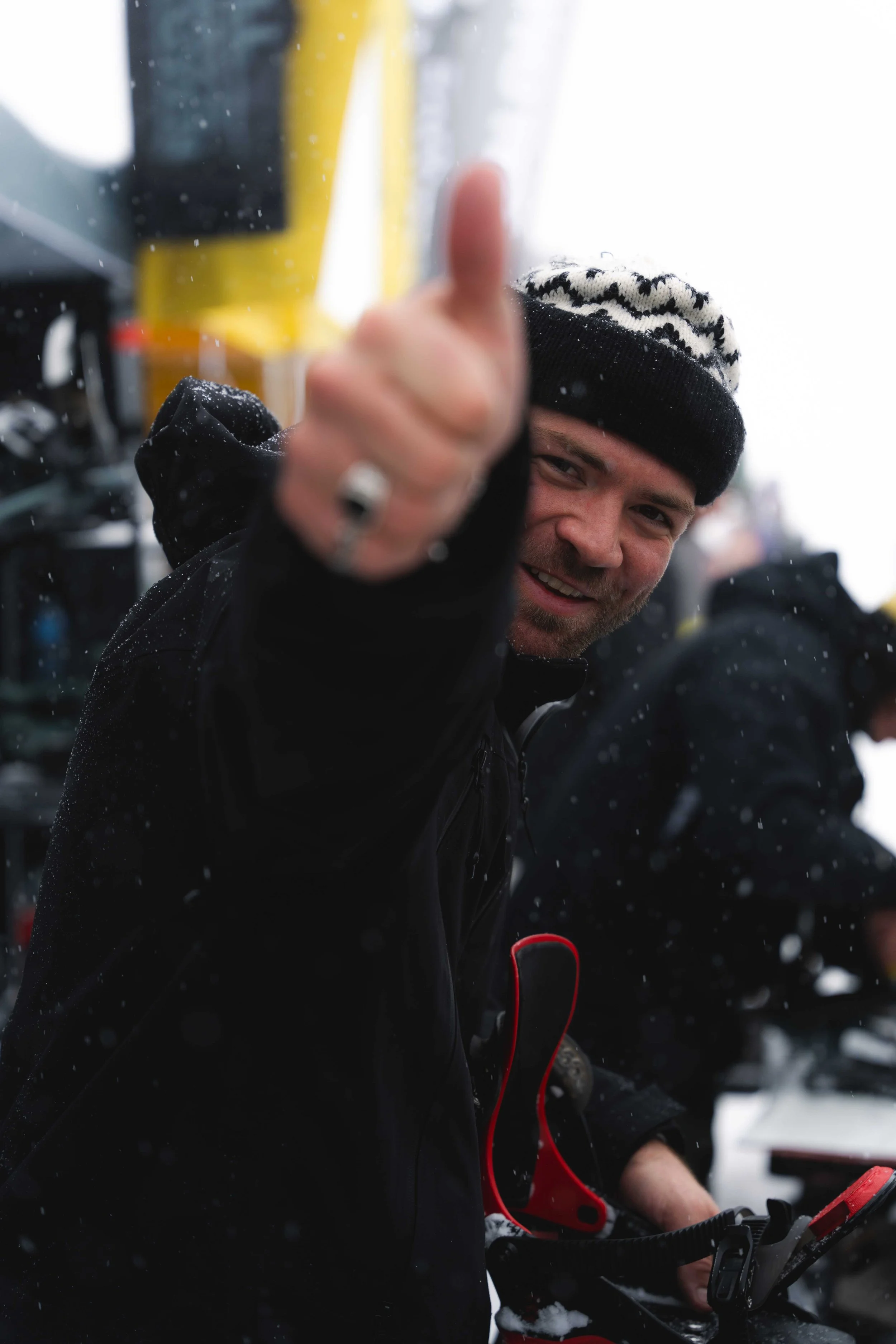 Homme souriant portant un bonnet noir et blanc, levant le pouce vers la caméra, avec fond enneigé.