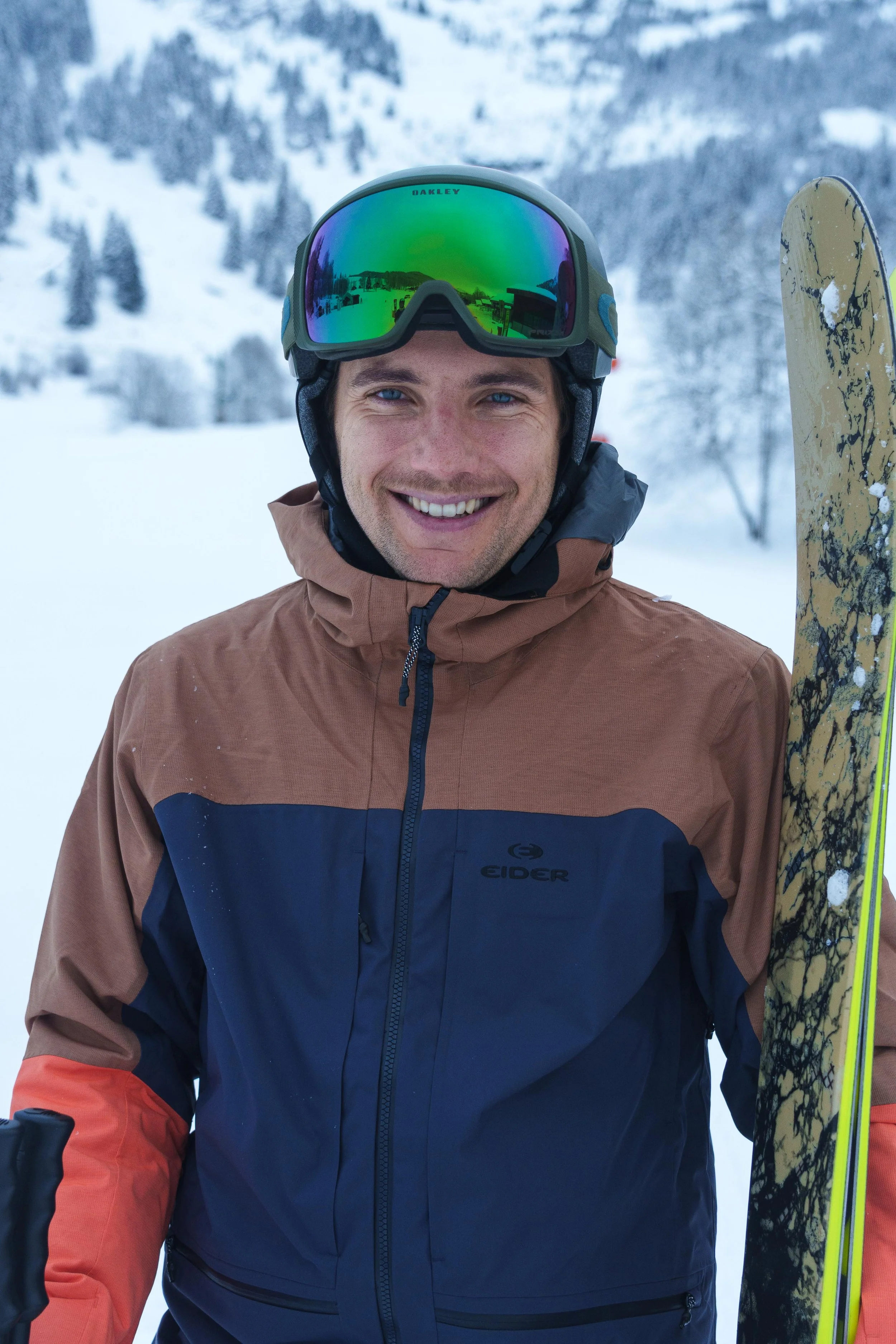 Skieur souriant avec des lunettes de ski et un casque, tenant des skis sur une piste enneigée.