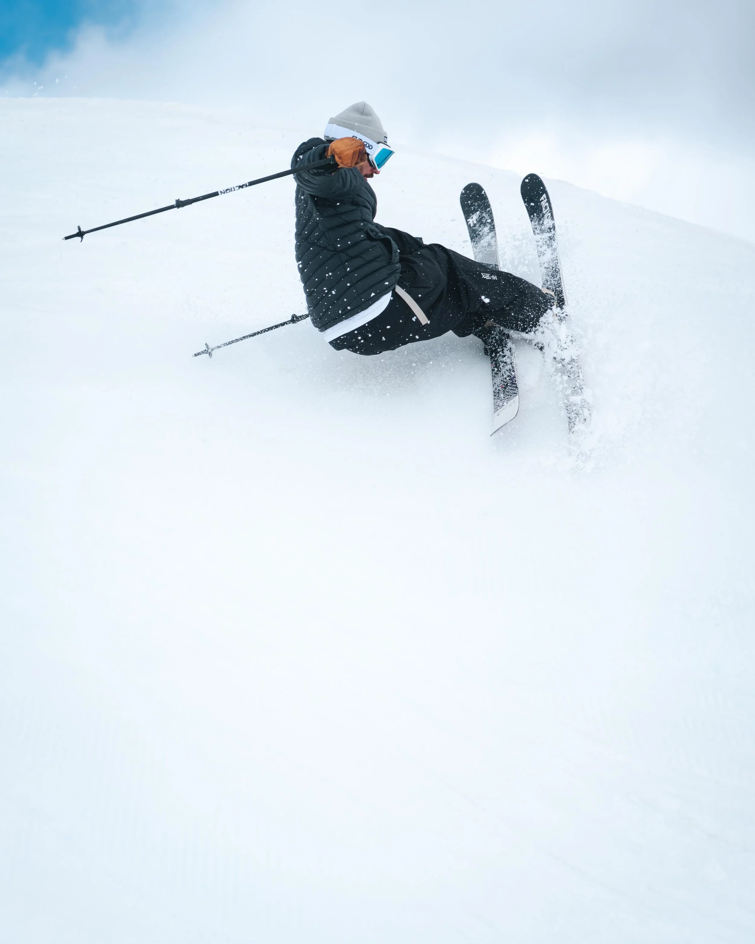 Personne faisant du ski dans la neige, portant une veste noire et un bonnet gris, utilisant des bâtons de ski, avec des skis parallèles et soulevant de la neige.