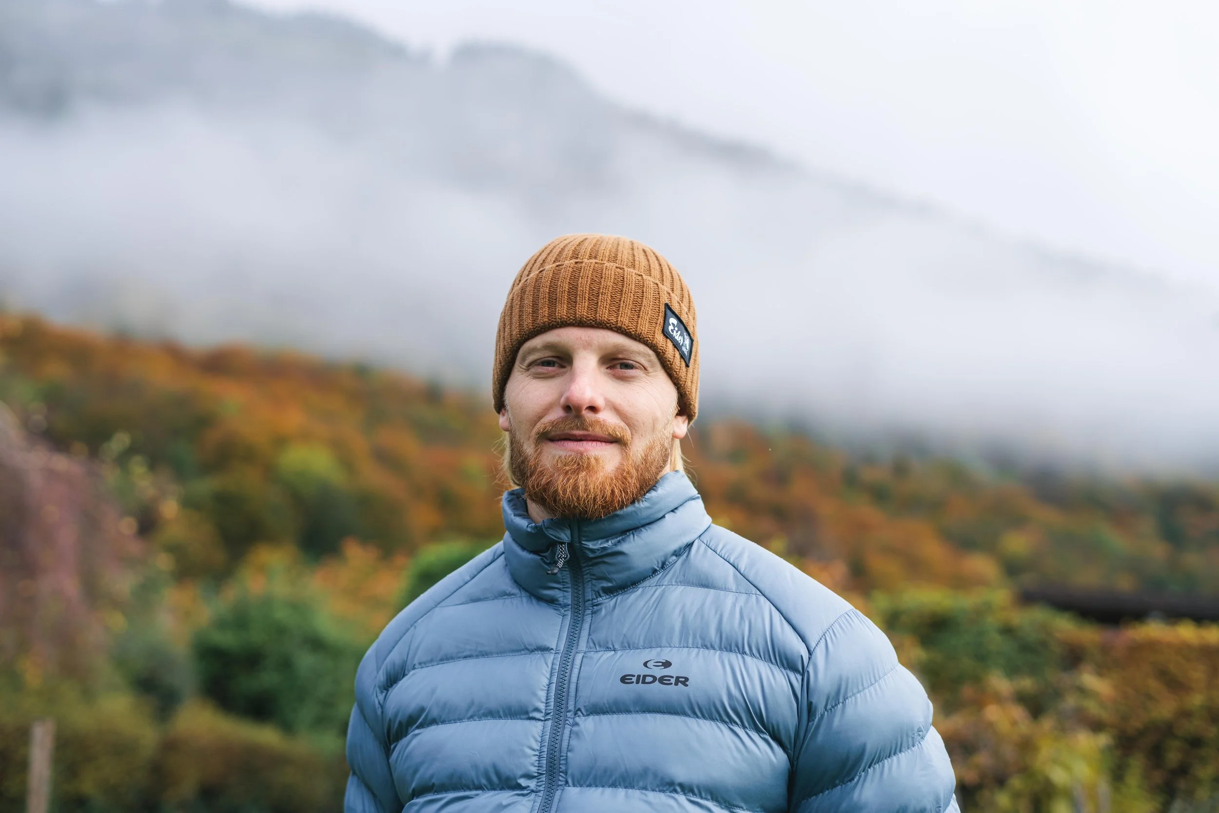 Homme portant une veste bleue et un bonnet marron devant un paysage automnal.