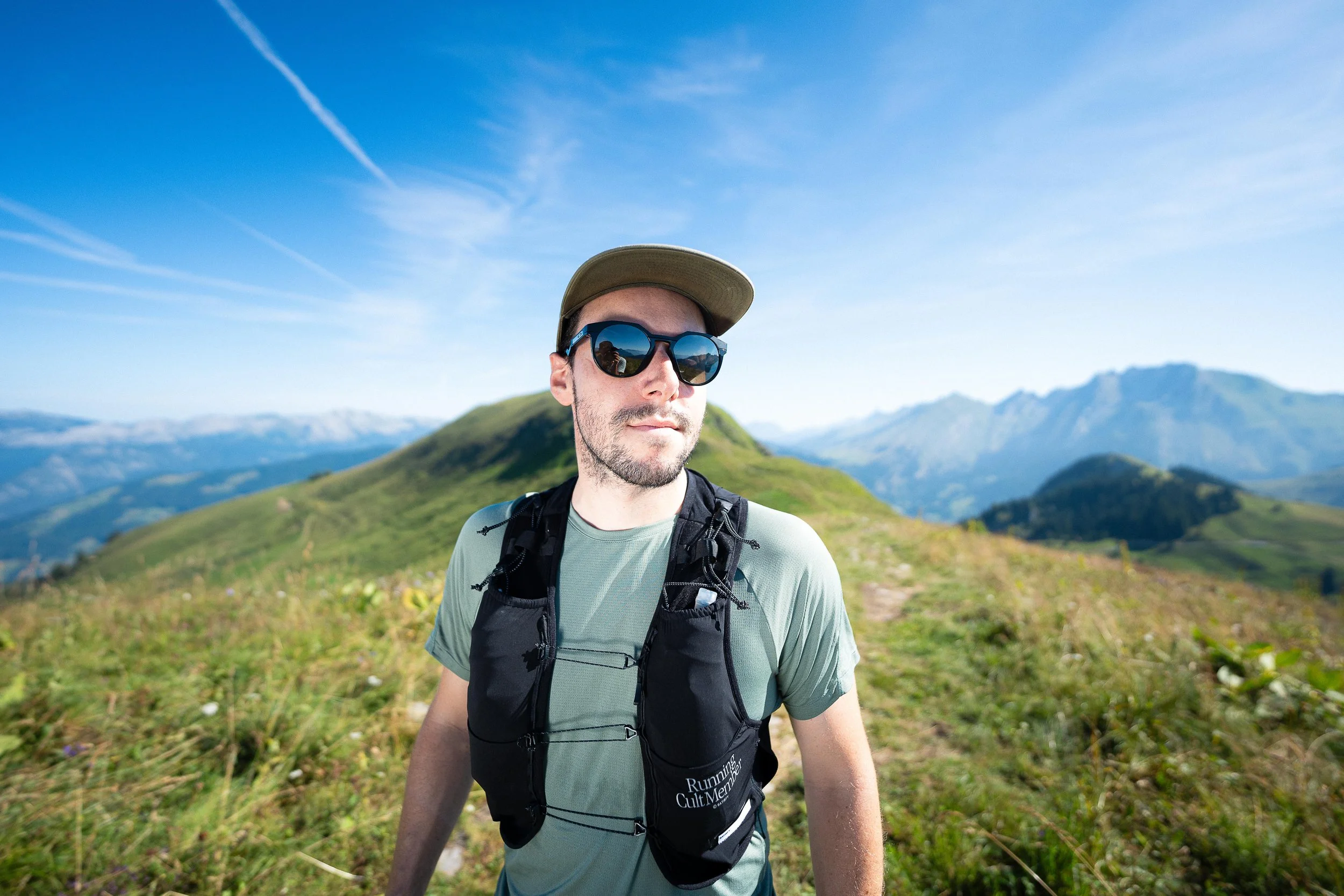 Un homme portant des lunettes de soleil et une casquette marche en plein air dans un paysage de montagne verdoyant sous un ciel bleu.