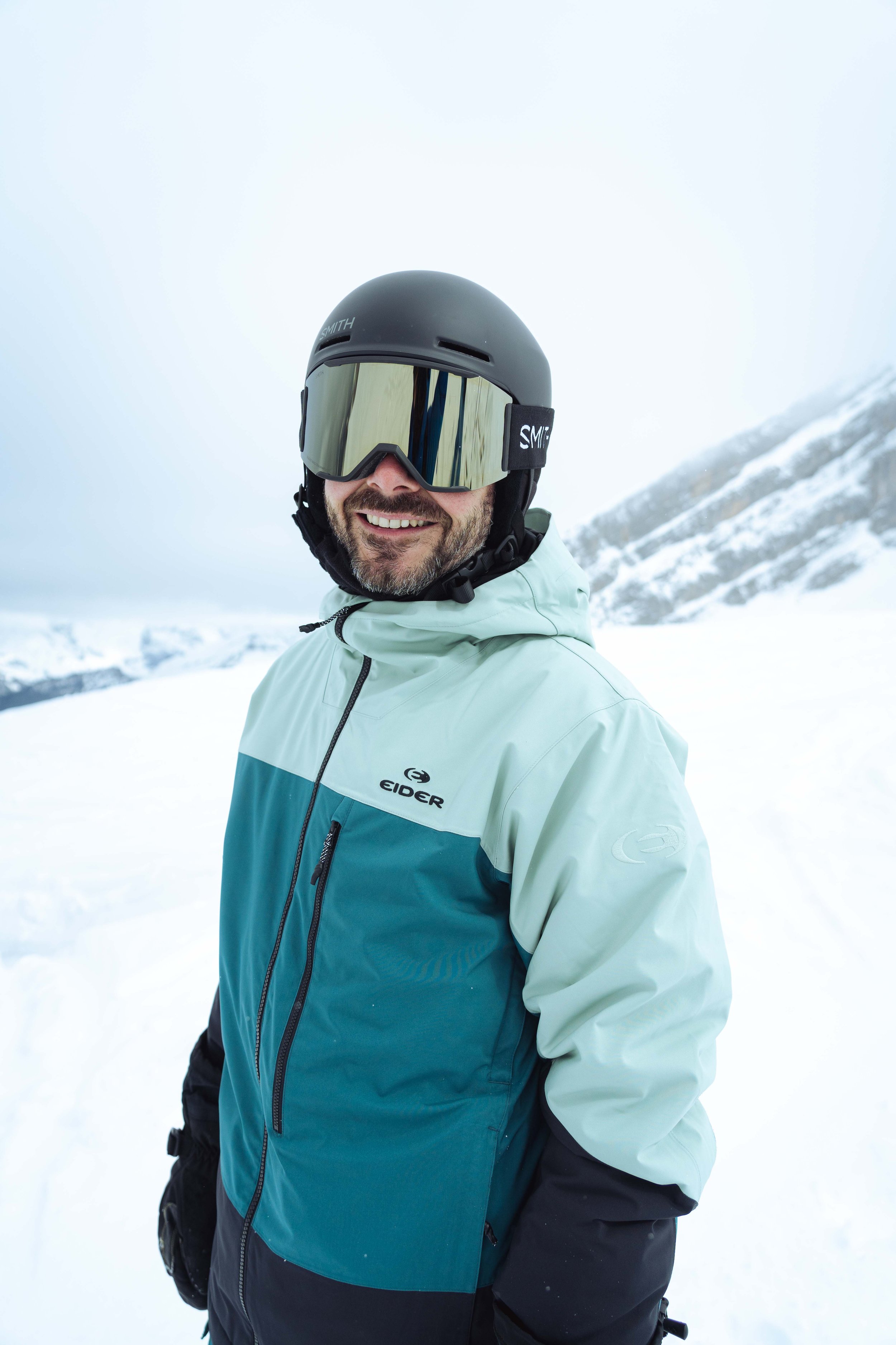 Personne souriant en ski, portant un casque et des lunettes, vêtu d'une veste de ski turquoise, sur une piste enneigée.