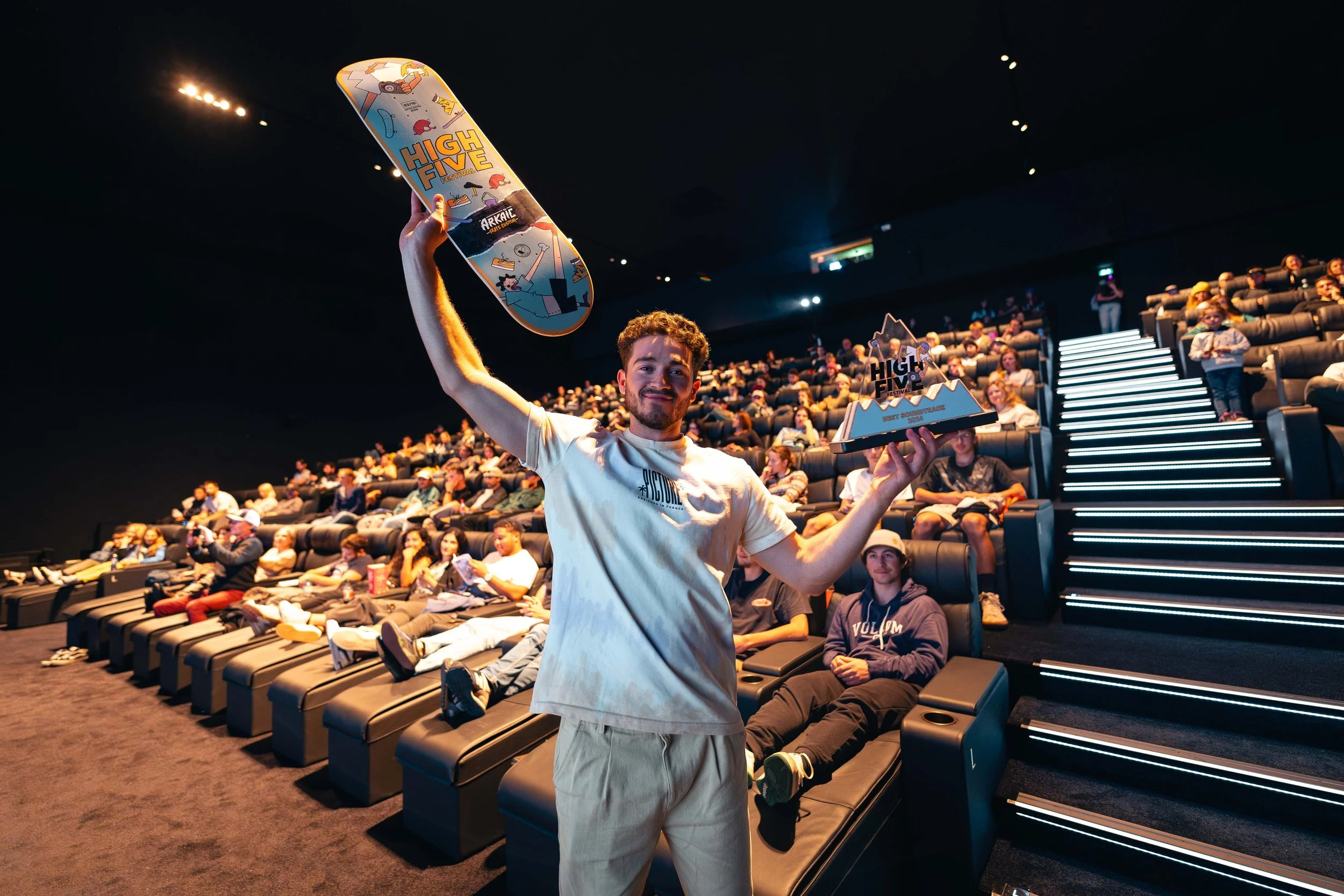 Un homme dans une salle de cinéma tenant un skateboard et un trophée "High Five", avec un public assis en arrière-plan.