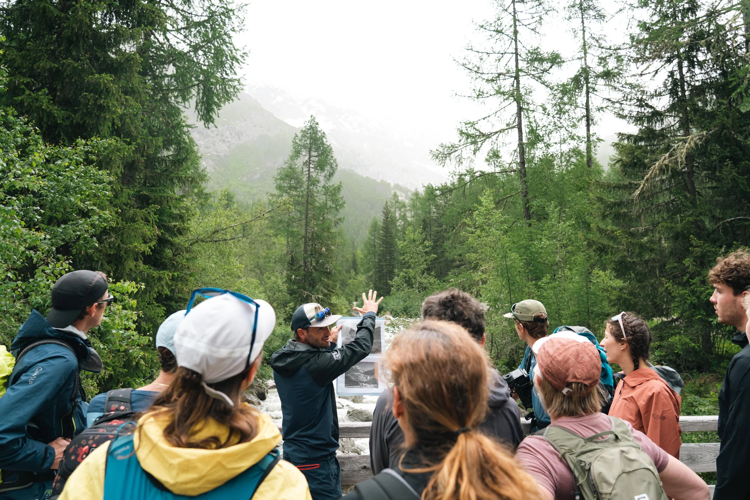Groupe de personnes lors d'une excursion en montagne, discutant avec un guide, entouré de forêts et de montagnes verdoyantes.