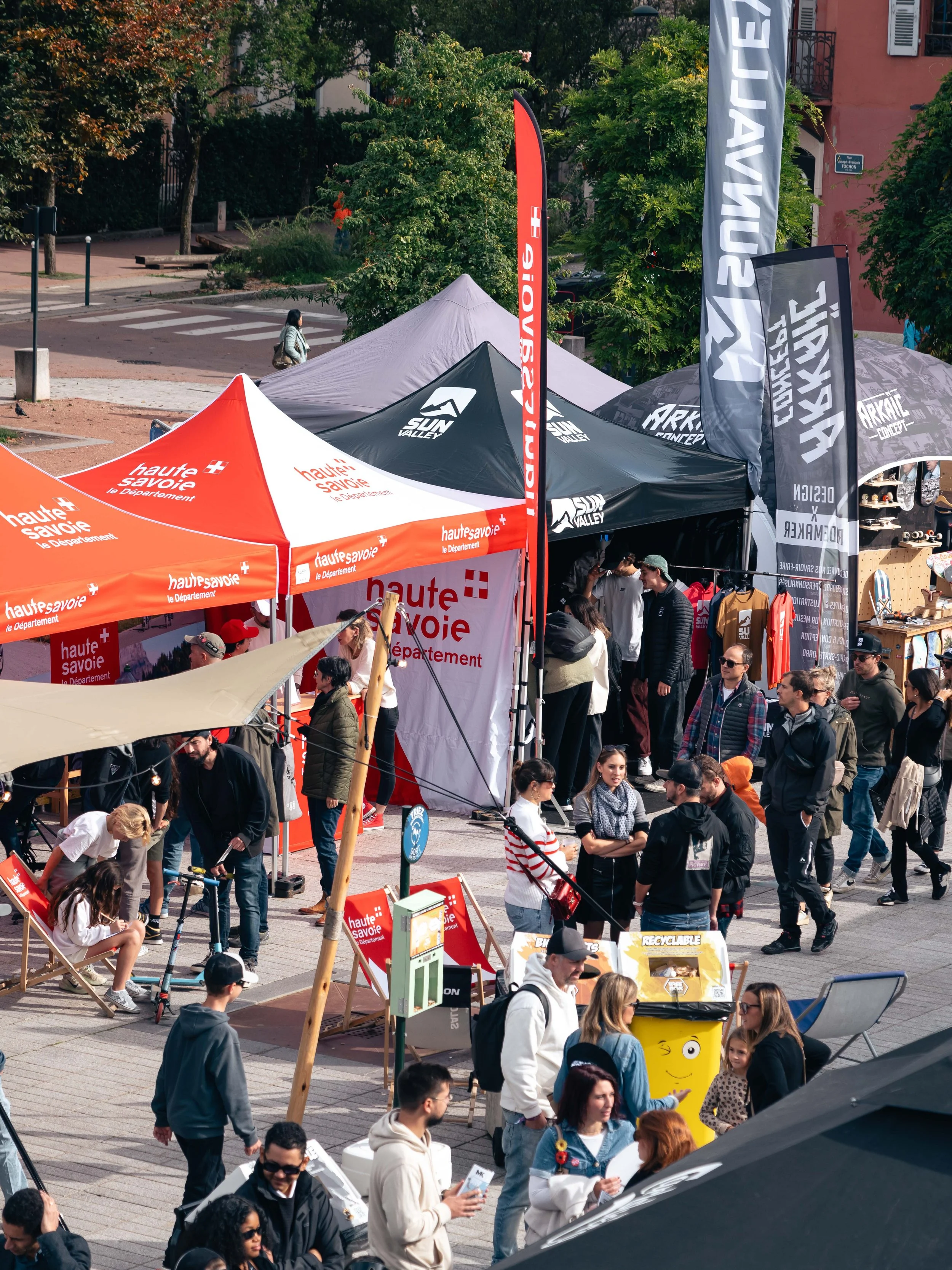 Un marché en plein air avec des stands de différentes marques, des gens se promenant sur la place, et des drapeaux indiquant "haute savoie" et "sun valley". L'ambiance est animée sous un ciel dégagé.