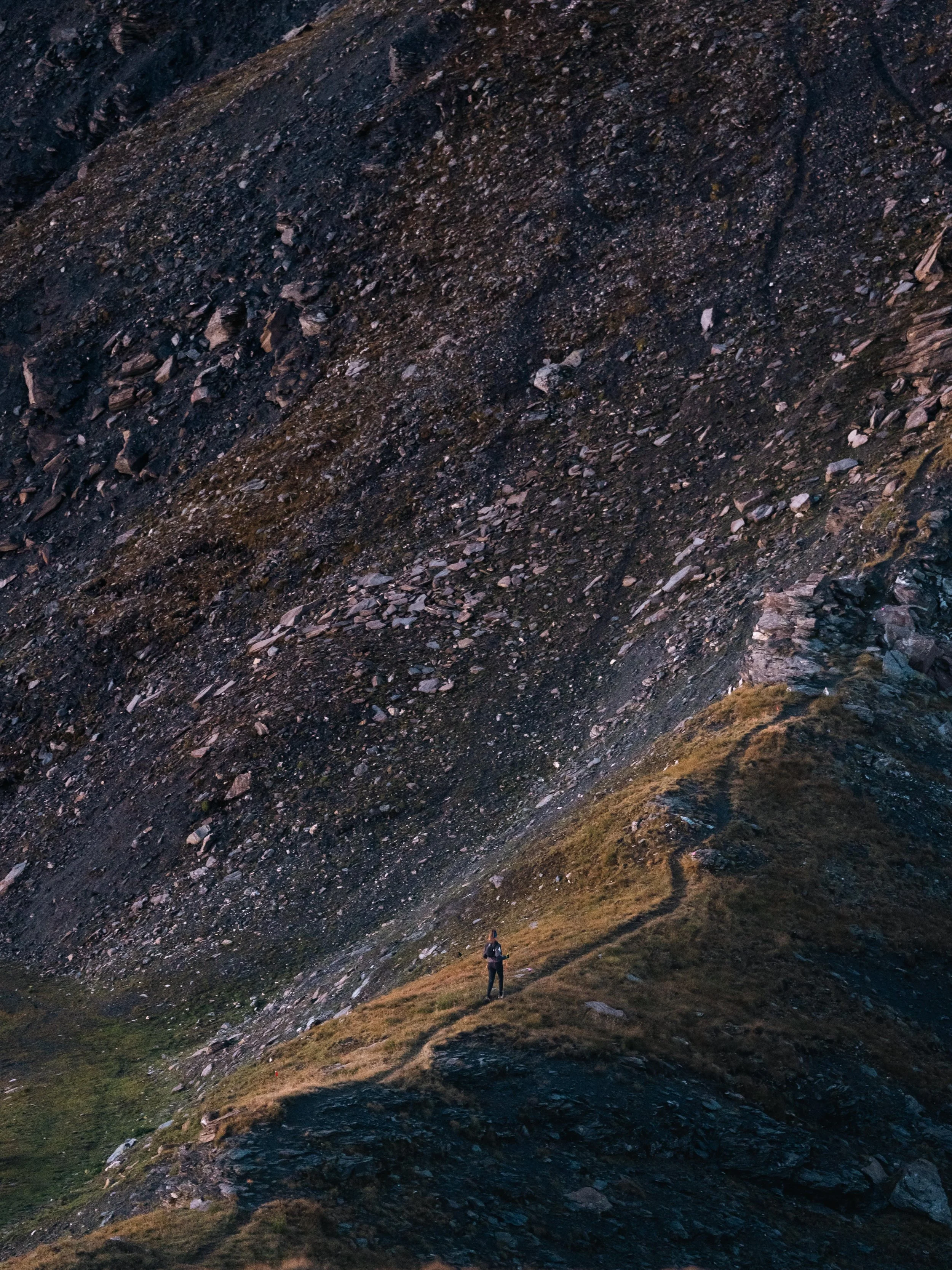 Paysage montagneux avec une personne sur un sentier herbeux, entourée de pentes rocailleuses.