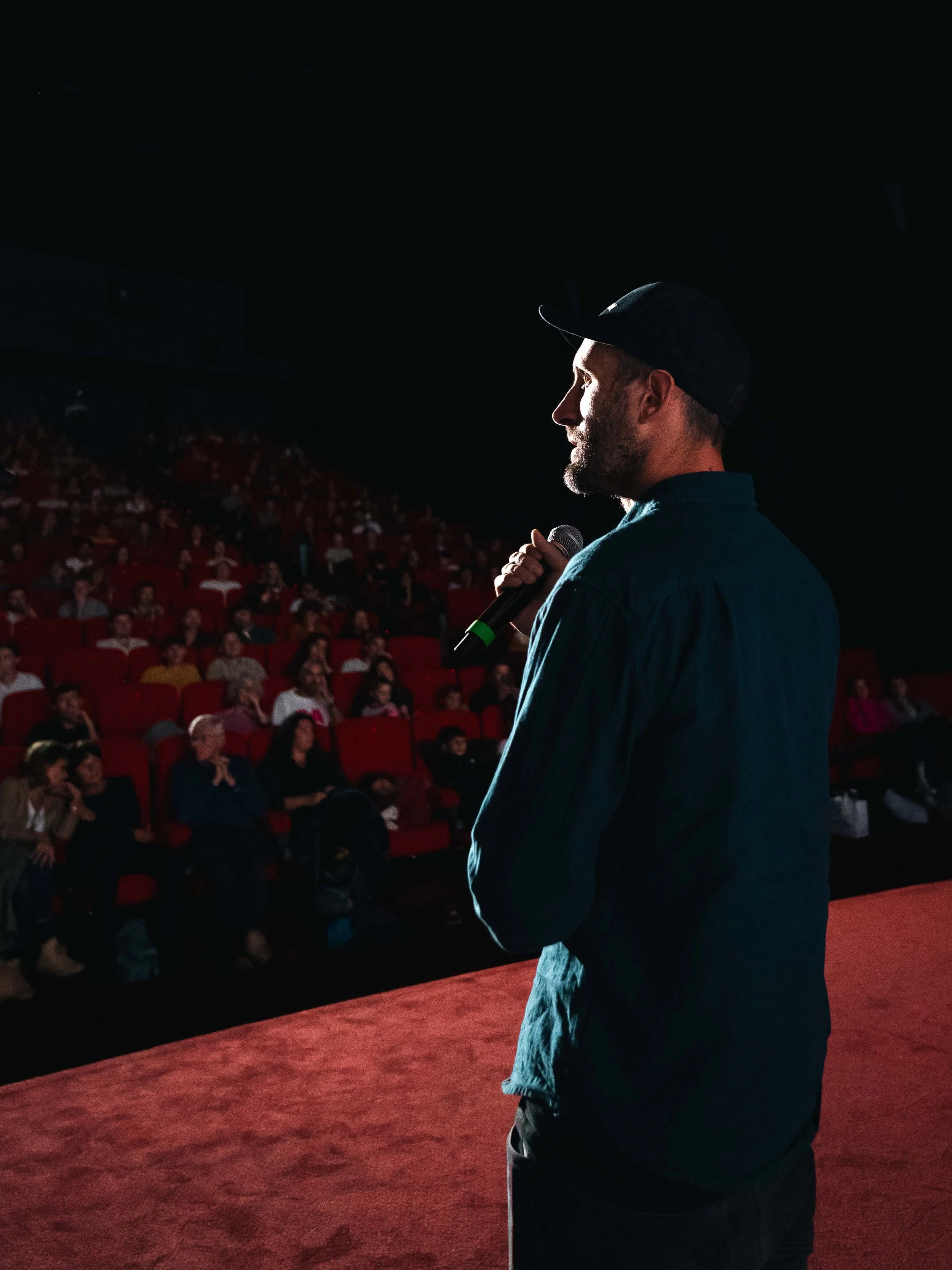 Un homme sur scène tenant un micro, s'adressant à un public assis dans un auditorium.