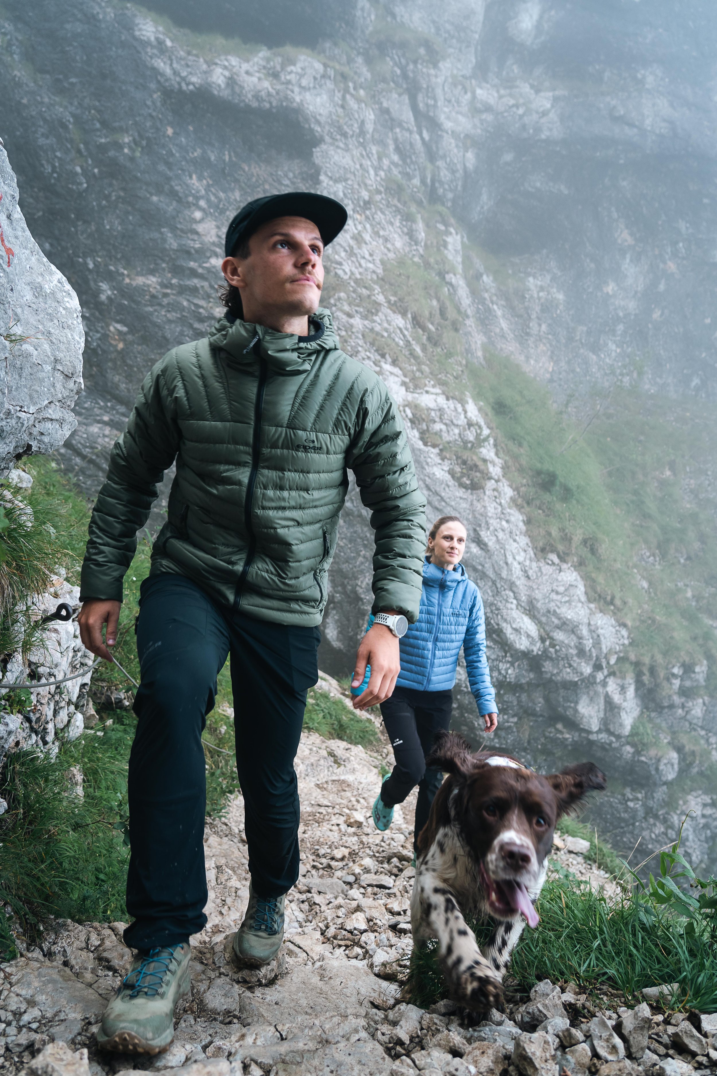 Un homme avec un blouson vert randonnée sur un sentier rocheux, accompagné d'une femme en blouson bleu et d'un chien à poil long. Le paysage est montagneux et brumeux.