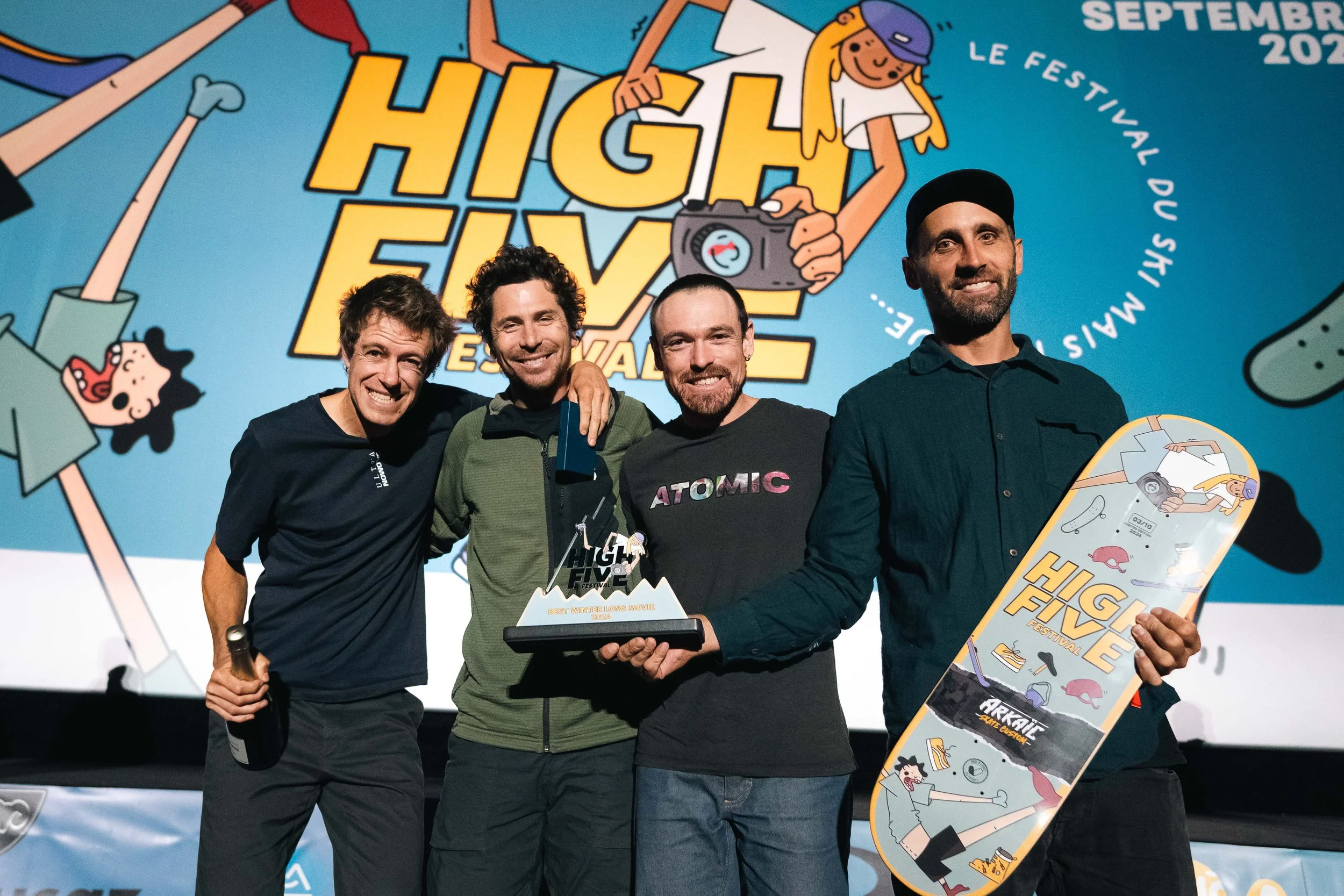 Un groupe de quatre personnes souriant sur scène lors du High Five Festival. Deux d'entre eux tiennent des trophées, dont un est en forme de montagne avec "High Five" inscrit dessus. En arrière-plan, une affiche colorée avec des illustrations et le t