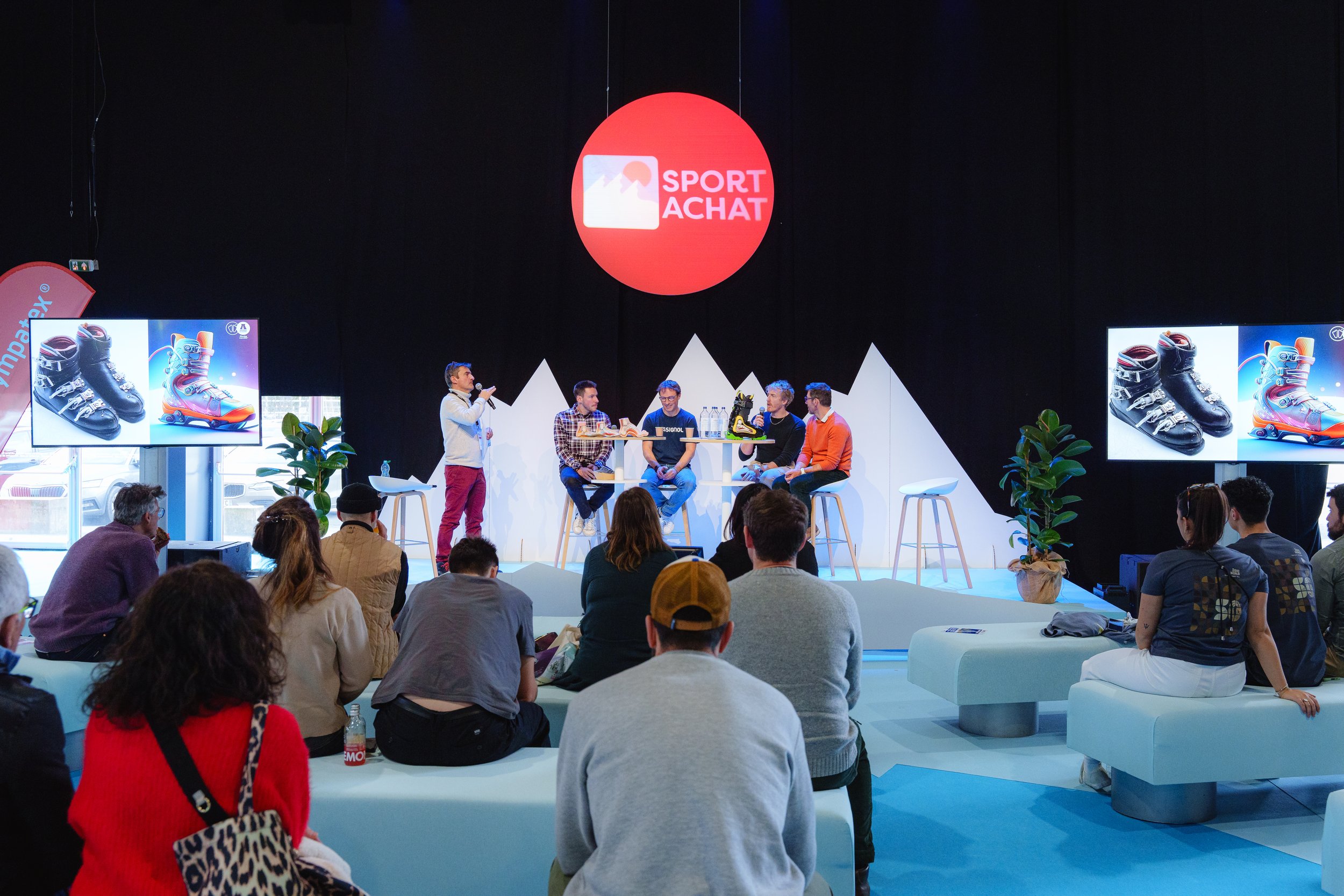 Une conférence se déroule dans un espace avec un grand logo "Sport Achat" en arrière-plan. Plusieurs personnes sont assises à une table ronde discutant probablement de produits sportifs, comme des chaussures de ski affichées sur des écrans. Un public
