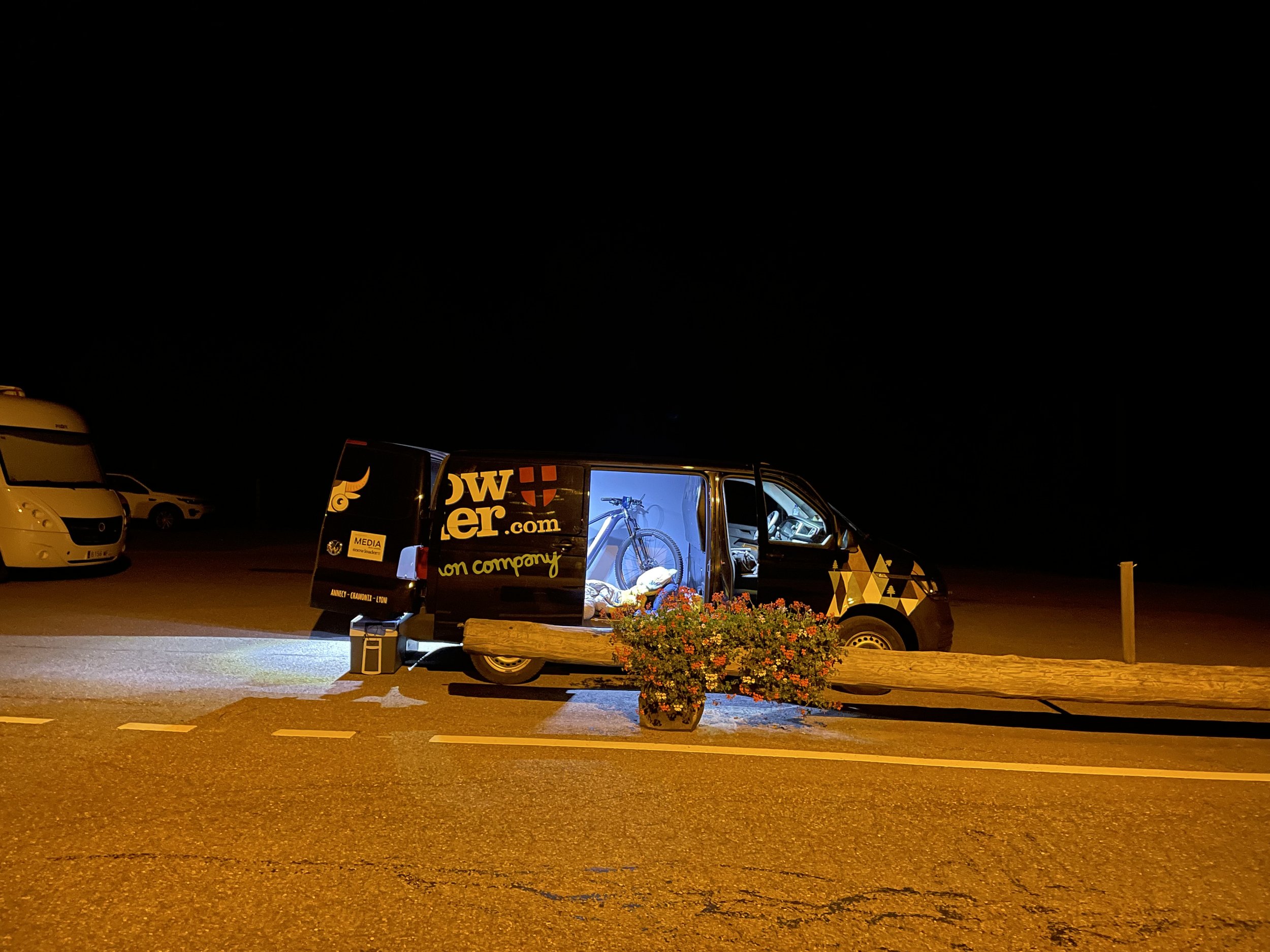Camionnette avec un vélo à l'intérieur, stationnée de nuit, portes ouvertes, lumière intérieure allumée, fleurs devant, véhicule et camping-car en arrière-plan.