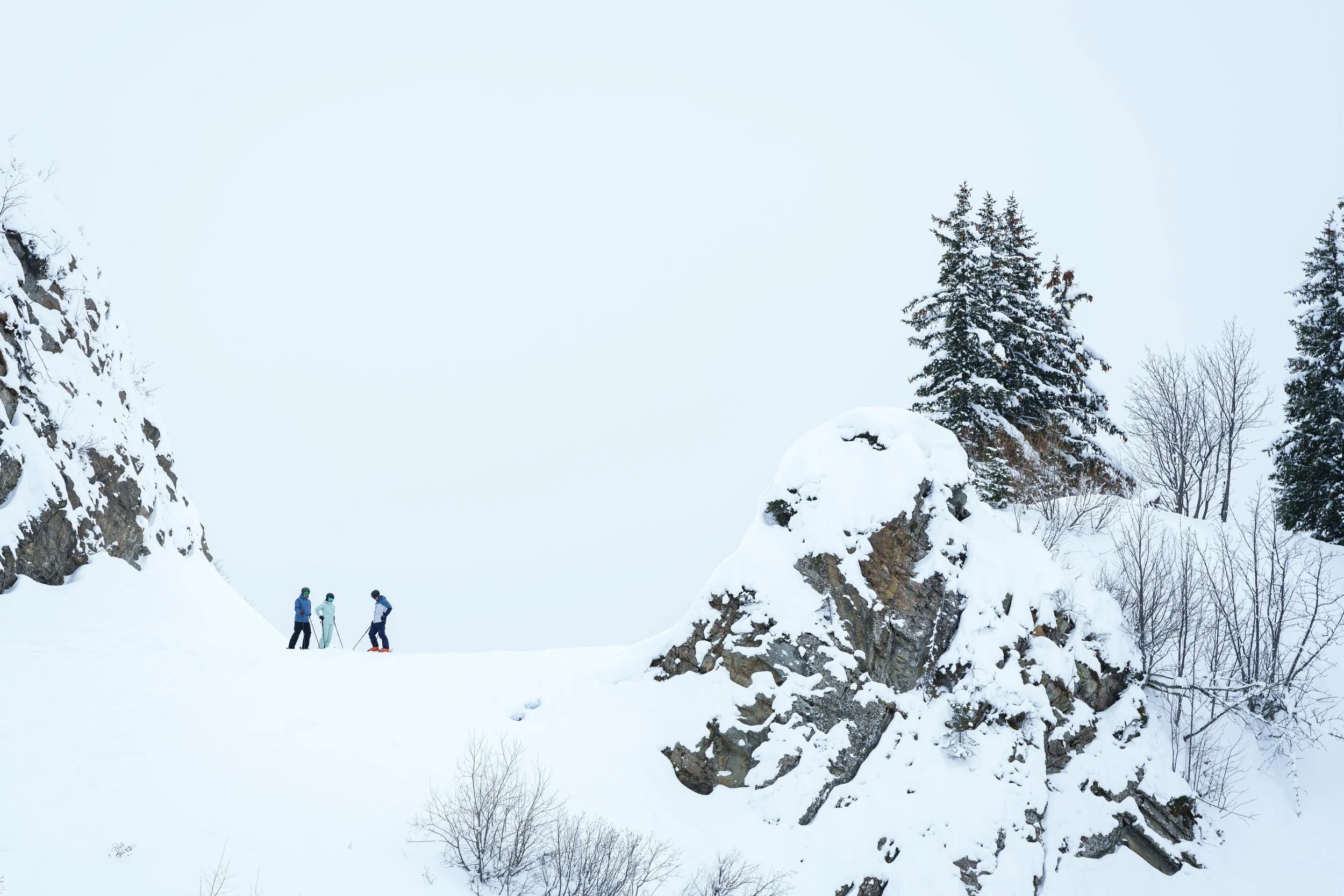 Trois personnes font du ski sur une montagne enneigée avec des sapins et des rochers.