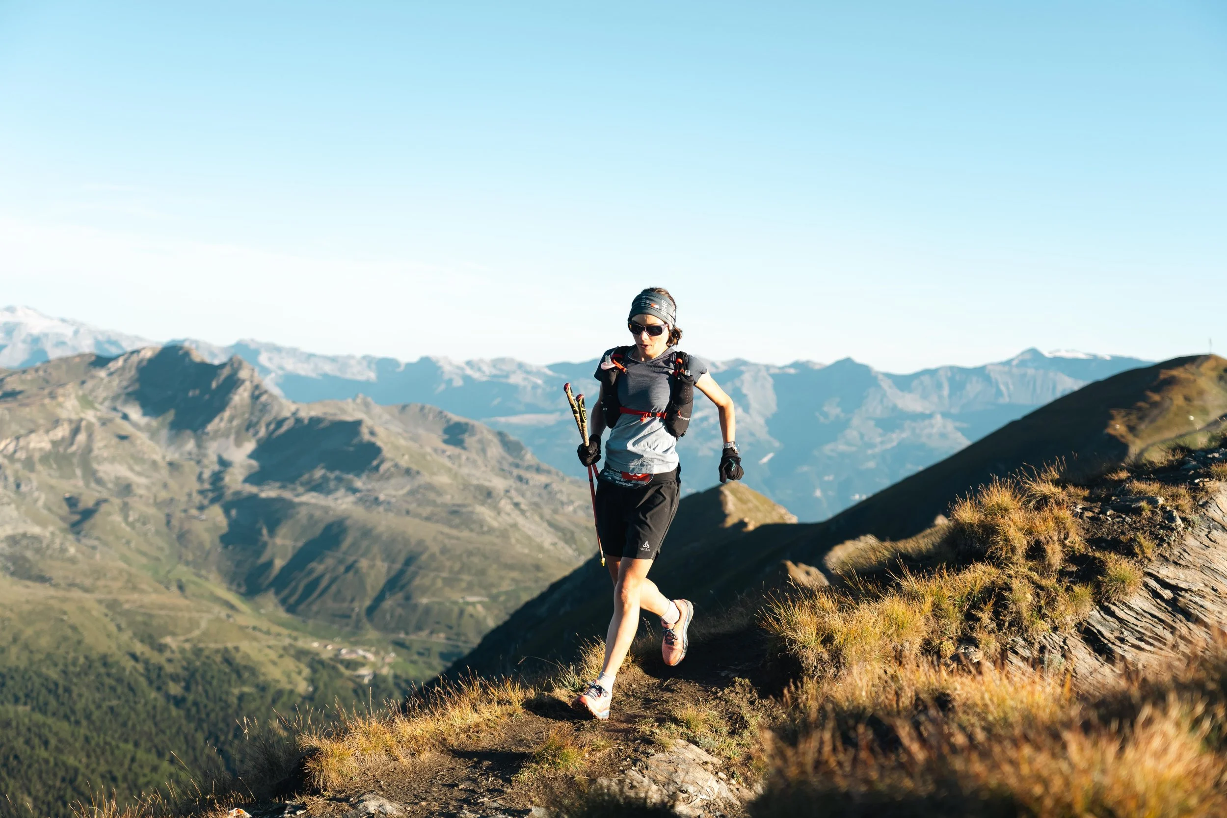 Une personne courant sur un sentier de montagne avec des montagnes en arrière-plan sous un ciel bleu.