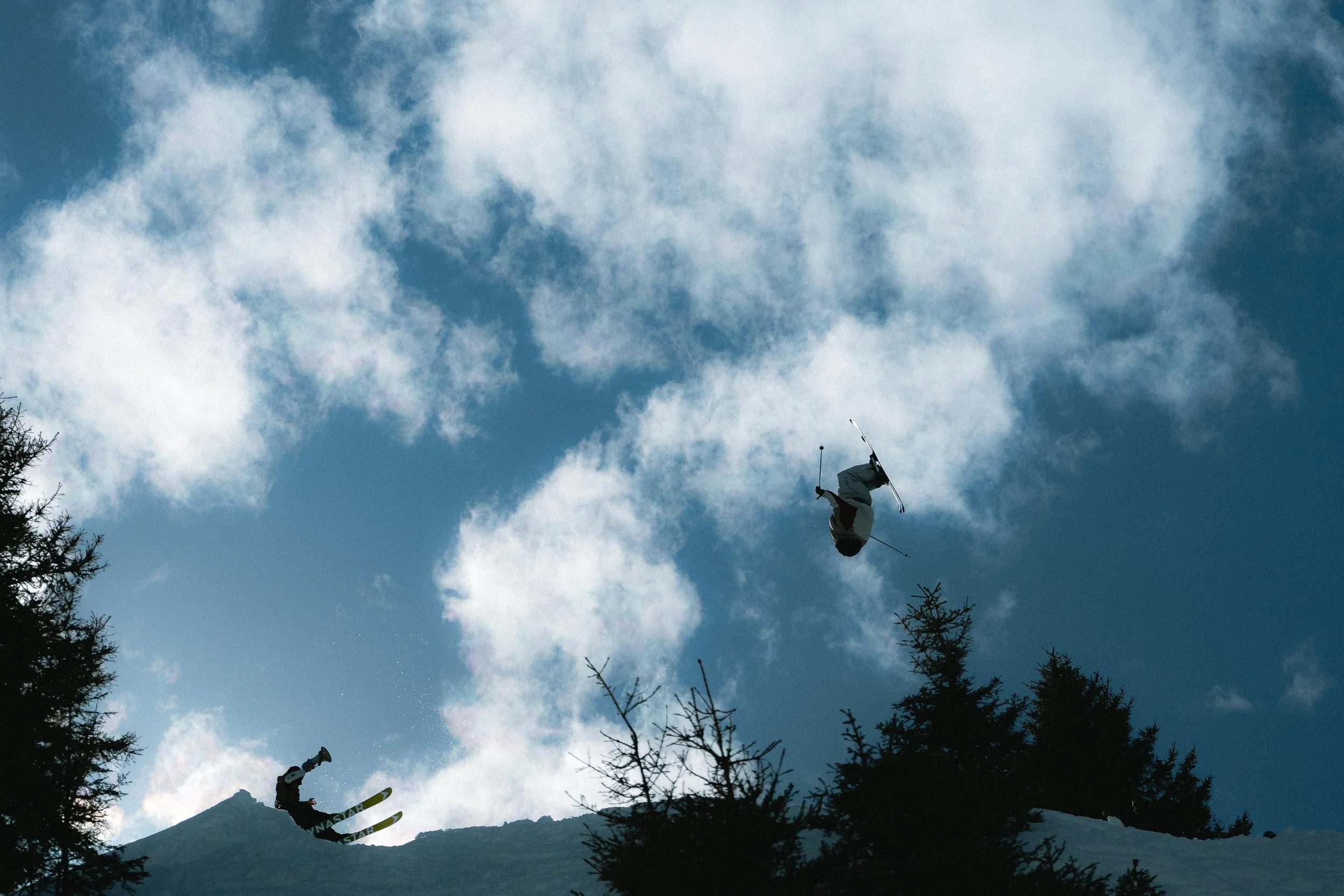 Deux skieurs effectuant des figures acrobatiques en l'air avec des arbres en contrebas et un ciel nuageux en arrière-plan. 
