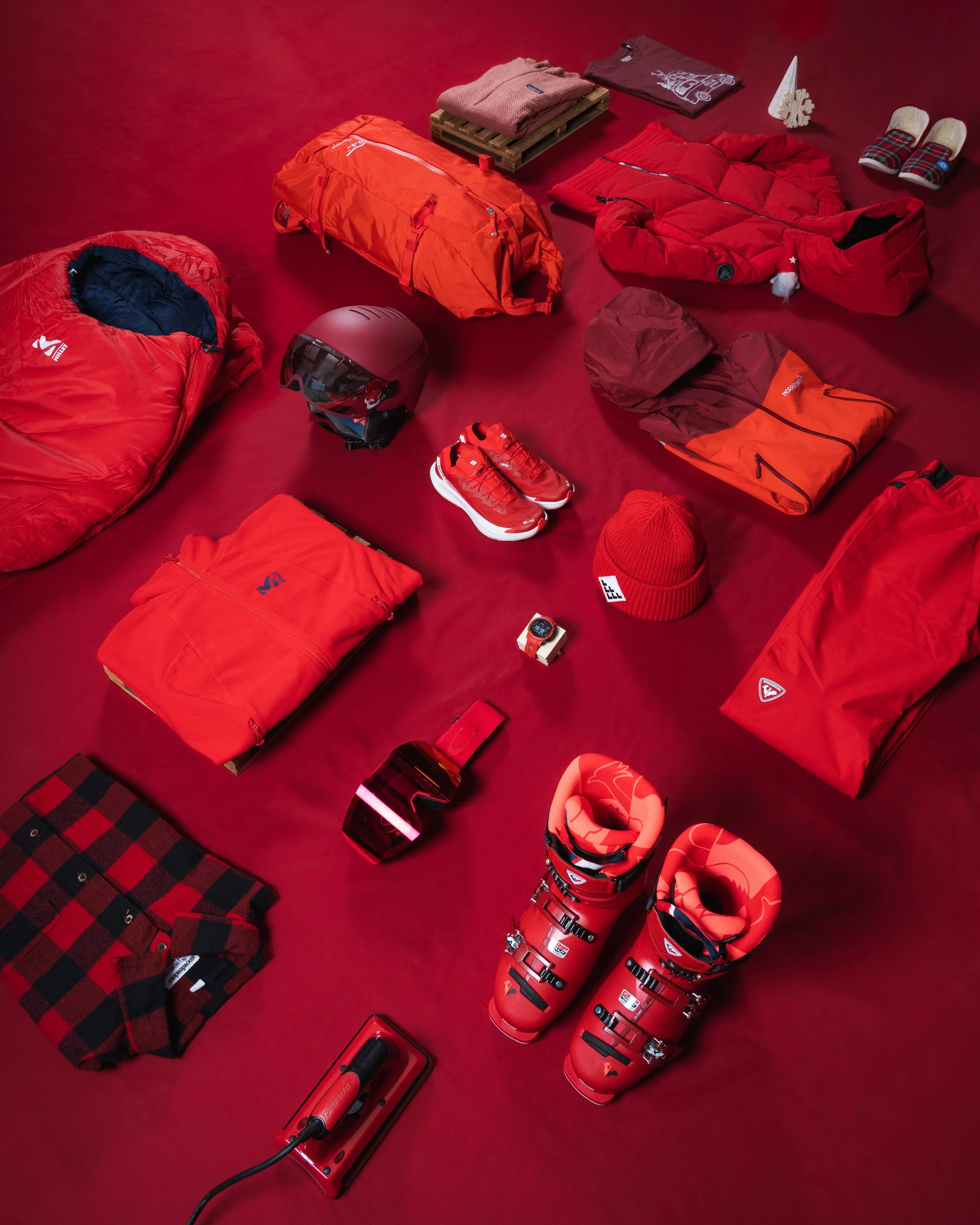 Vêtements et équipements sportifs rouges, dont un sac de couchage, un sac de voyage, une doudoune, un casque, des chaussures, des lunettes de ski, un bonnet et des gants, disposés sur un fond rouge.