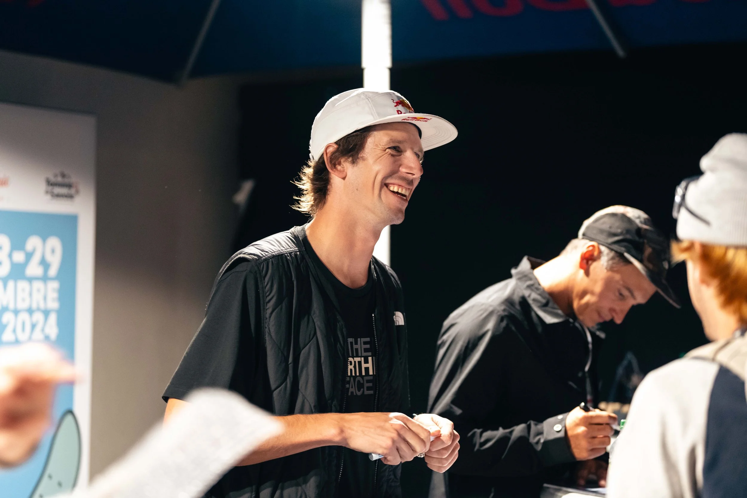 Deux hommes portant des casquettes, l'un souriant et discutant, l'autre écrivant, dans un événement public.