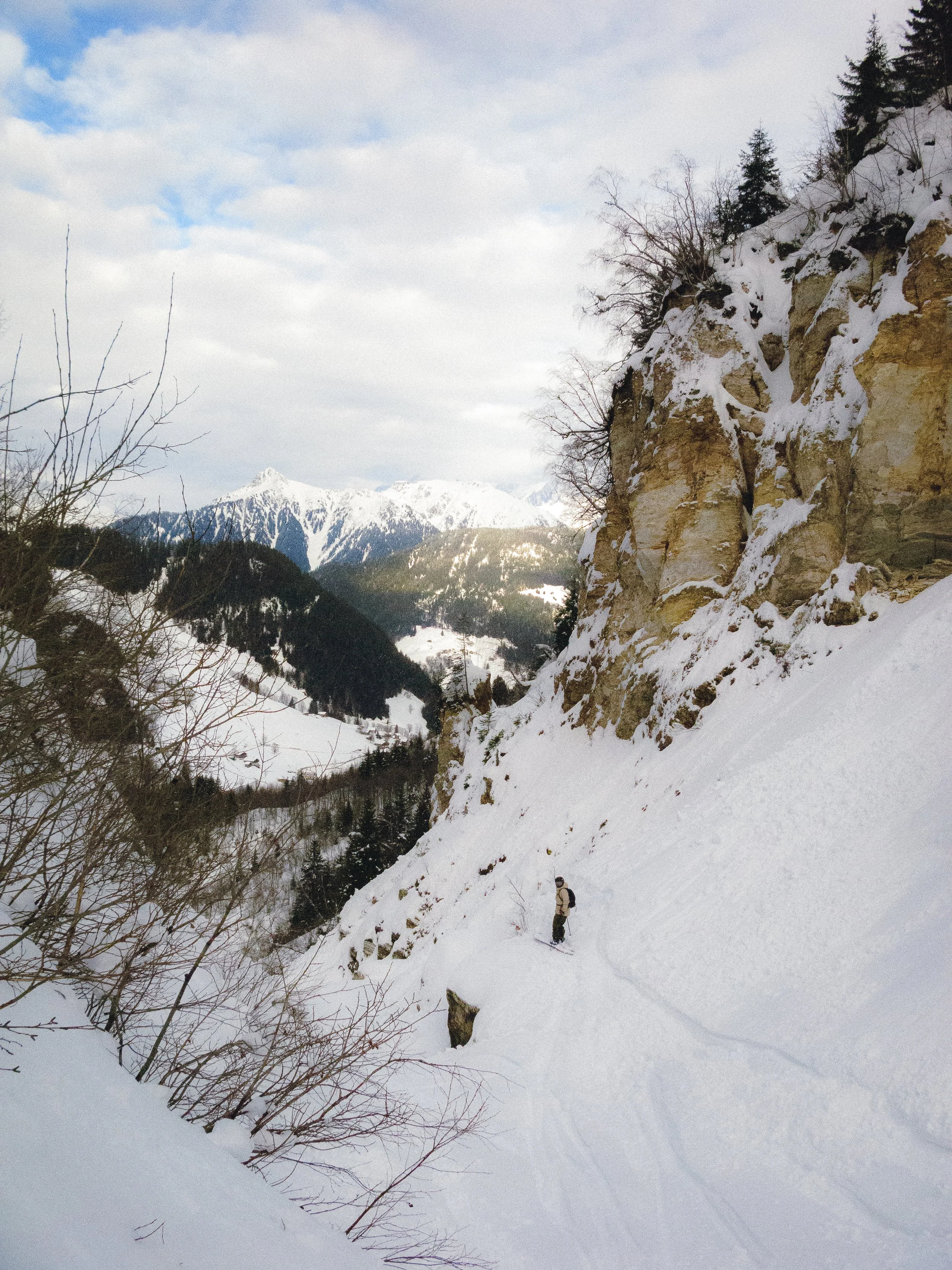 Paysage hivernal montagneux avec un skieur ou randonneur dans une vallée enneigée, arbres dénudés, ciel partiellement nuageux, montagnes en arrière-plan.