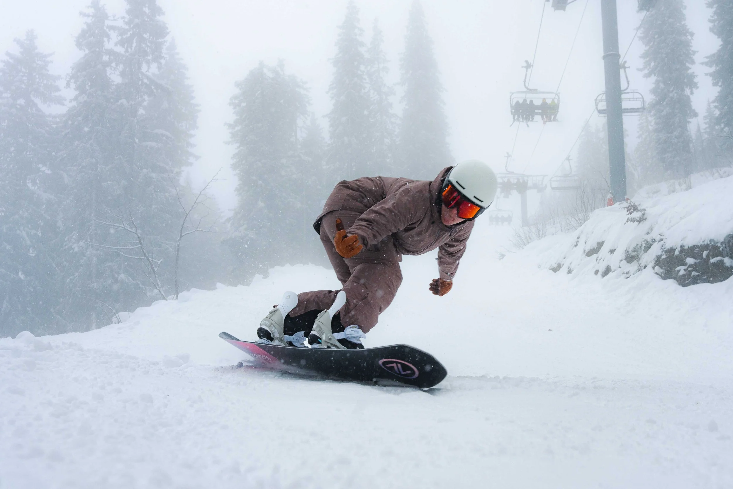 Une personne en snowboard sur une piste enneigée avec des arbres et un télésiège en arrière-plan.