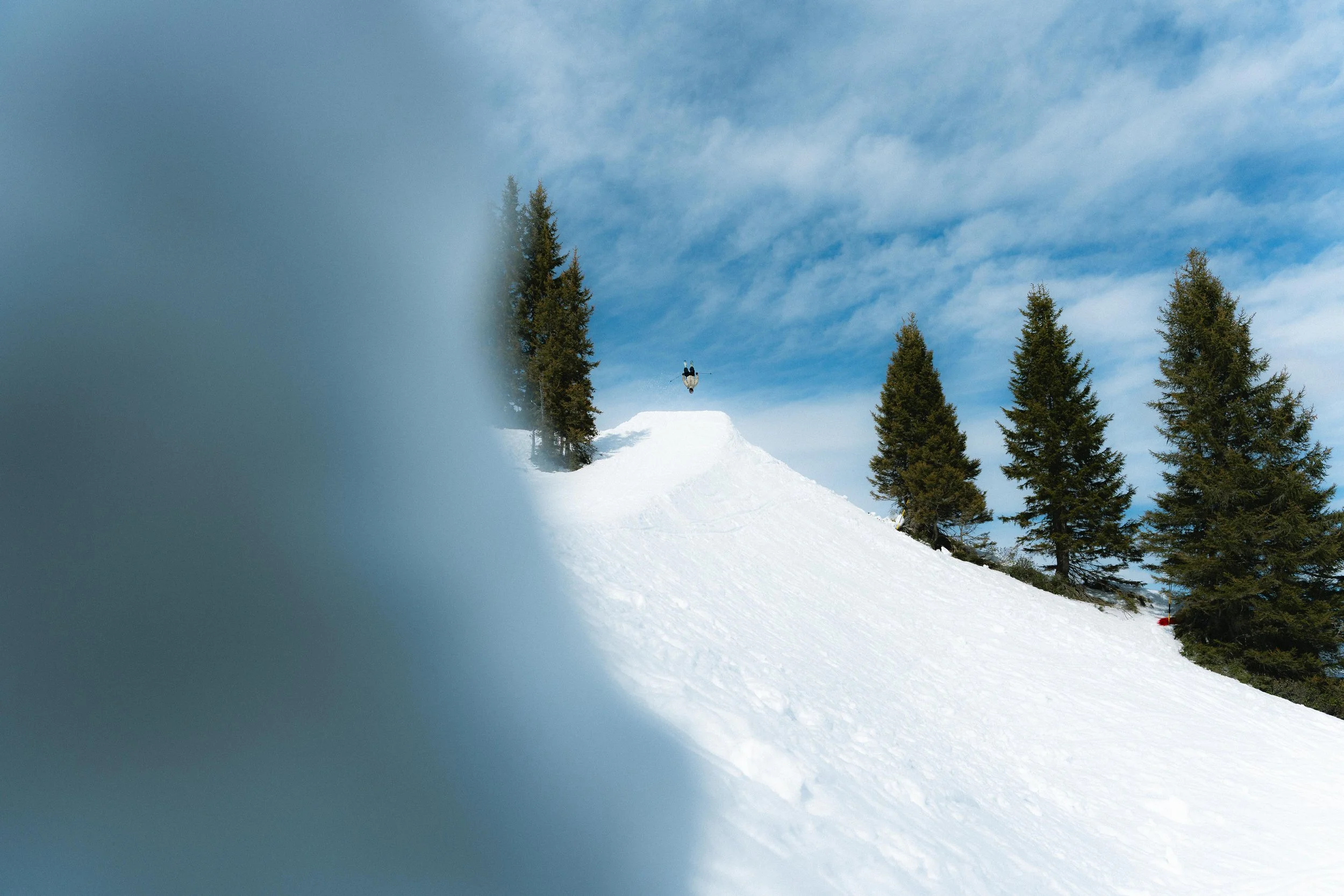 Un skieur effectue un saut sur une rampe enneigée avec des sapins en arrière-plan et un ciel bleu partiellement nuageux.