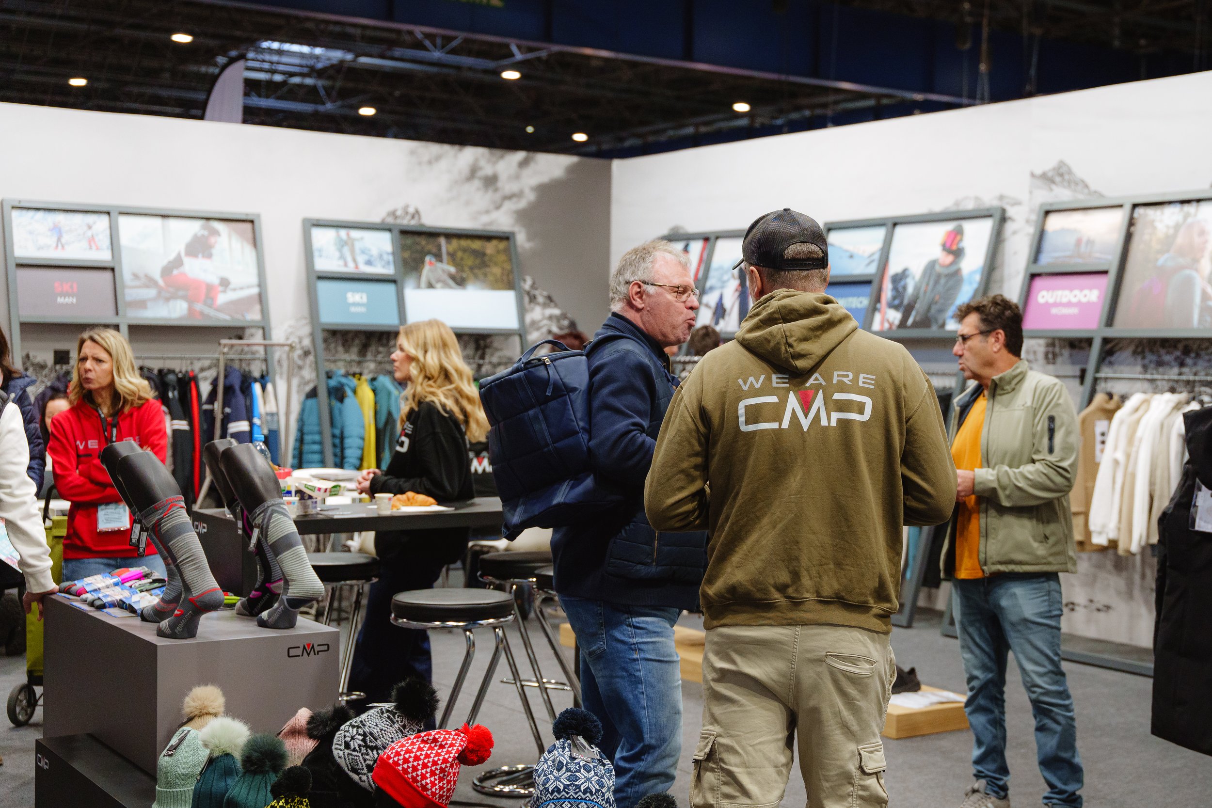 Un salon professionnel de vêtements de sports d'hiver, avec des gens discutant autour de stands de présentation, où sont exposés des manteaux et des accessoires comme des chaussettes et bonnets. Des écrans affichent des images et descriptions de prod