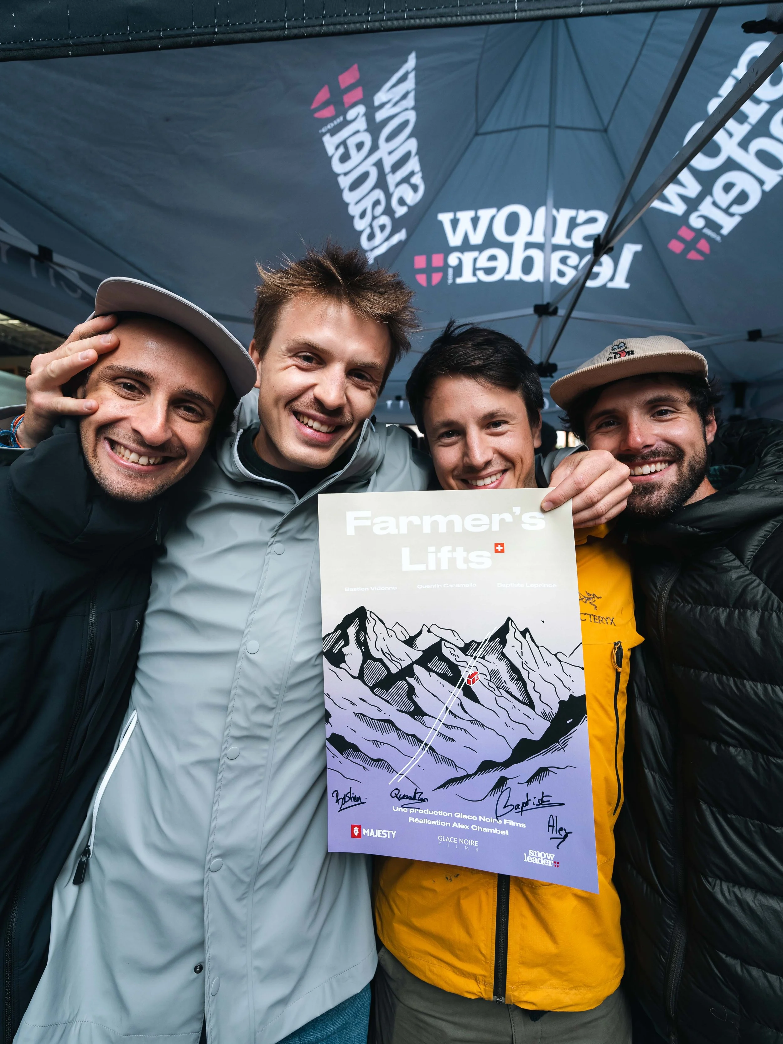 Quatre personnes souriantes posent ensemble sous une tente avec un panneau indiquant "Farmer's Lifts". Le fond montre un paysage montagneux dessiné. Ils portent des vestes et des chapeaux d'hiver.