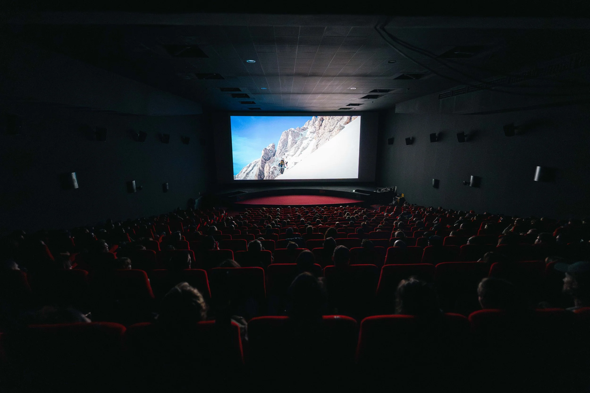 Salle de cinéma avec écran projetant un film de montagne, spectateurs assis sur des sièges rouges.