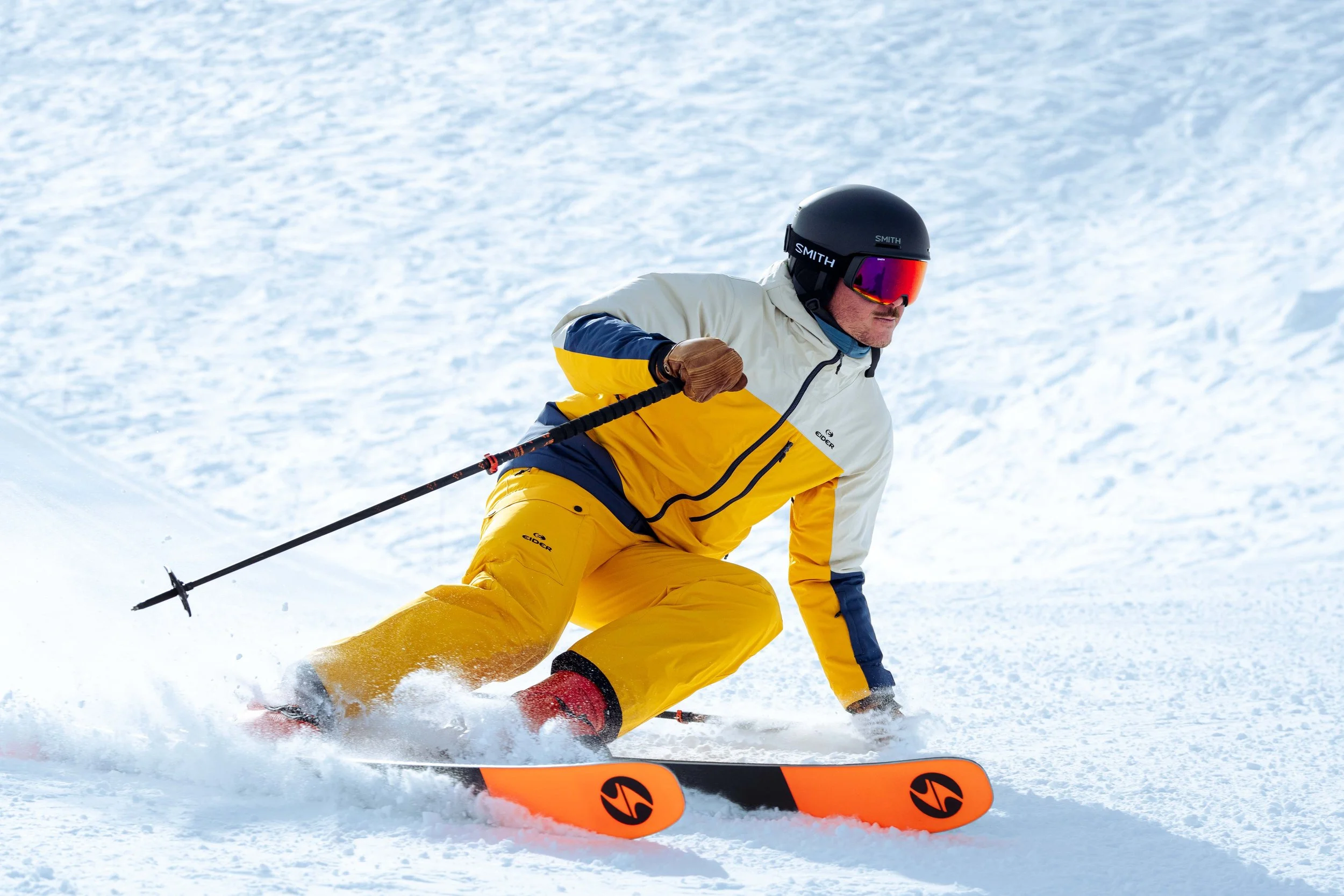 Une personne fait du ski sur une pente enneigée, vêtue d'une combinaison de ski jaune et blanche, avec un casque et des lunettes de soleil.