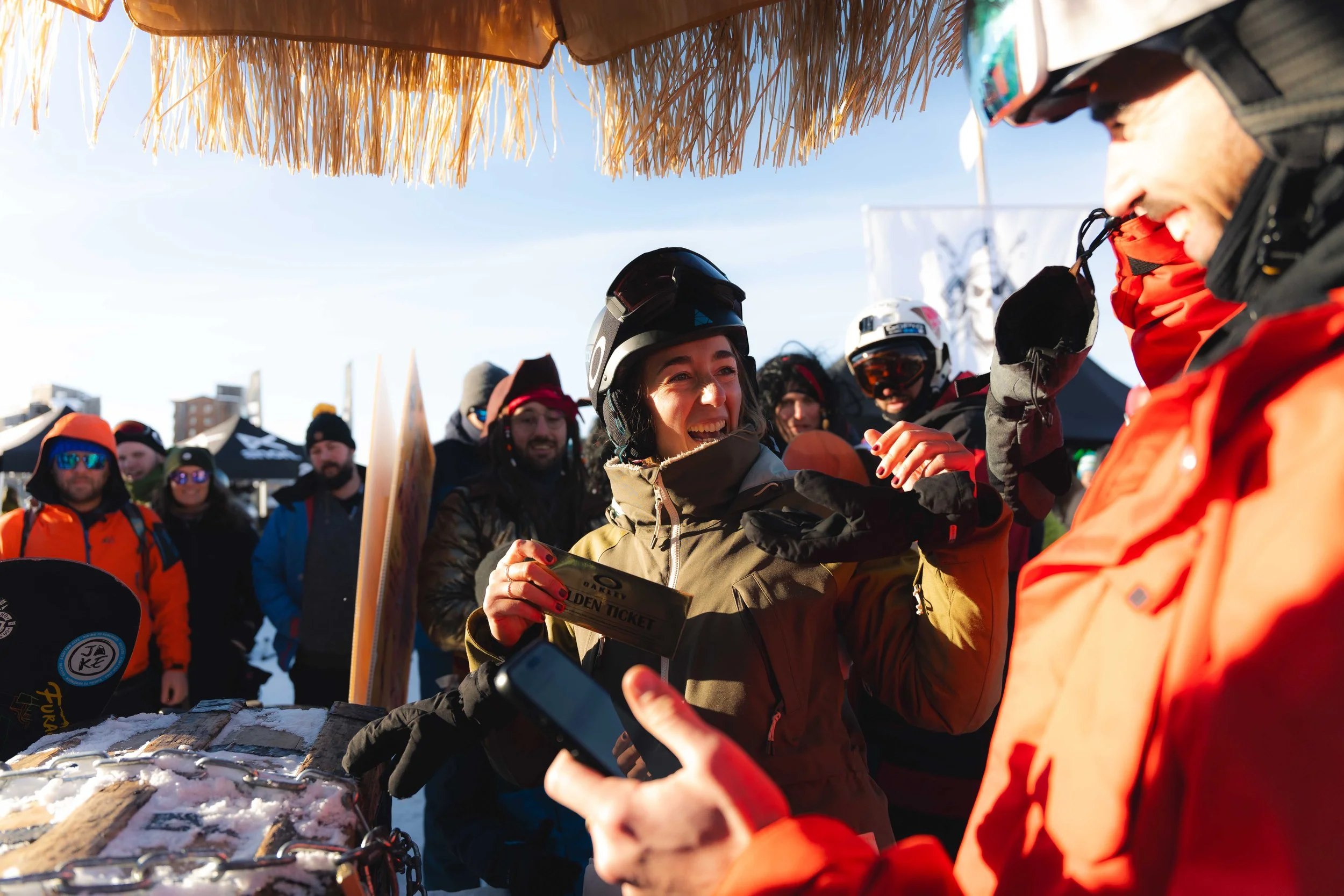 Personnes en vêtements de ski lors d'un événement en plein air, une femme tenant un billet doré, entourée de spectateurs, sous un parasol de paille.