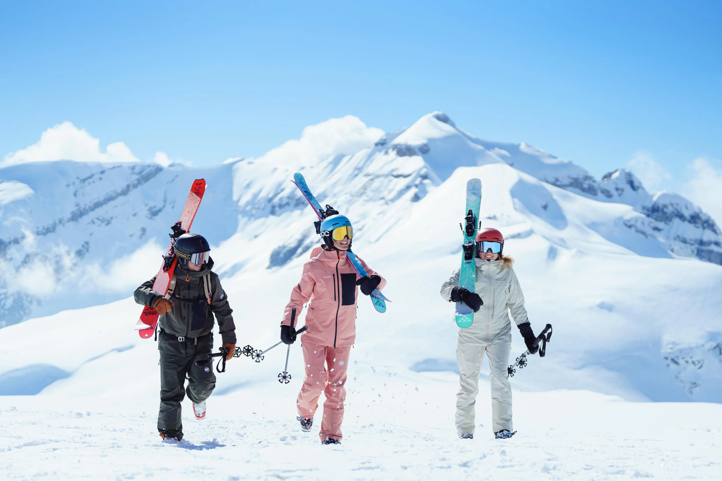 Trois skieurs marchant sur la neige portant leurs skis, avec des montagnes enneigées en arrière-plan.