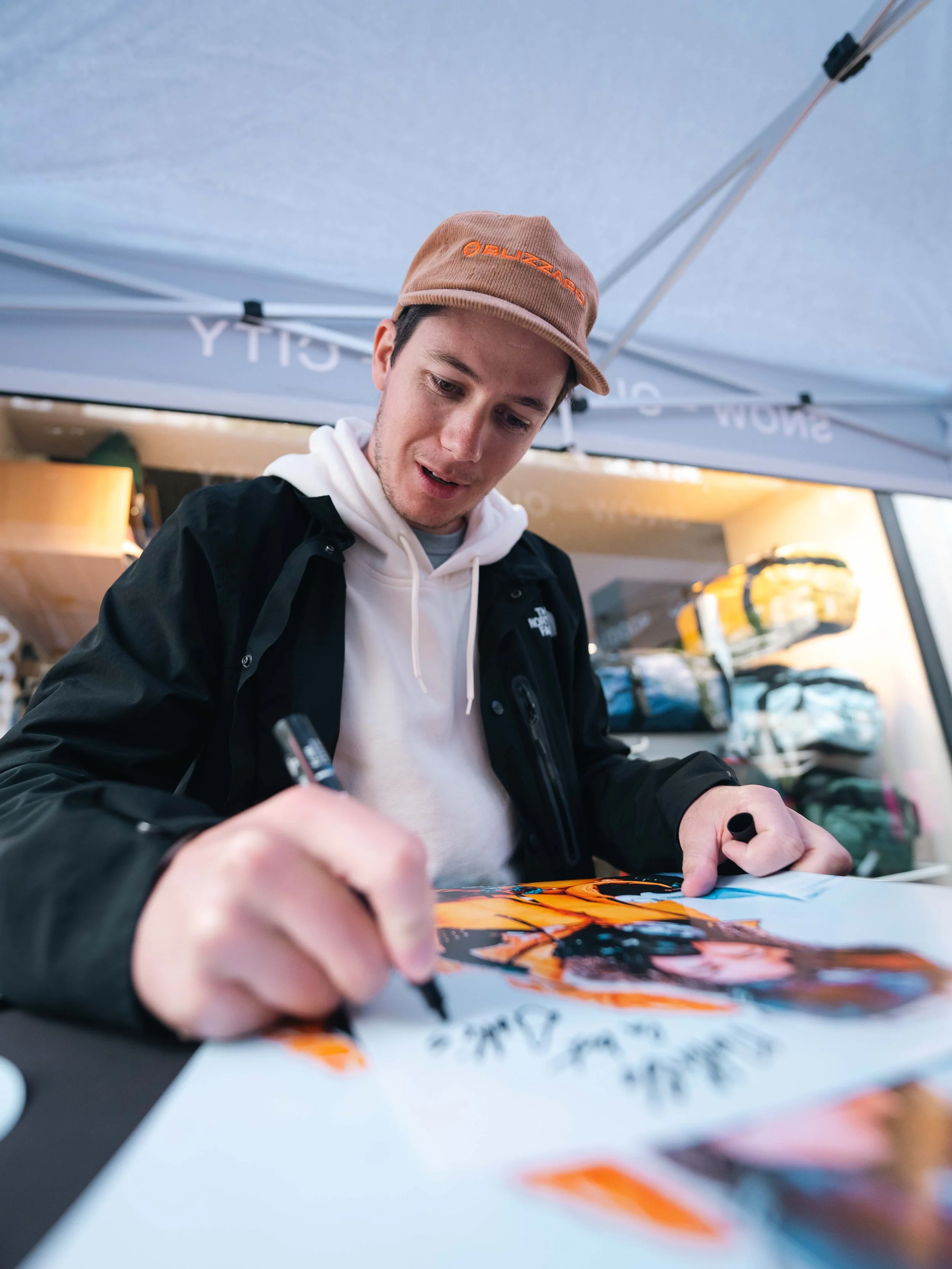 Un homme portant une casquette marron et un sweat à capuche blanc écrit sur une affiche colorée. Il se trouve sous une tente et semble concentré sur son travail.
