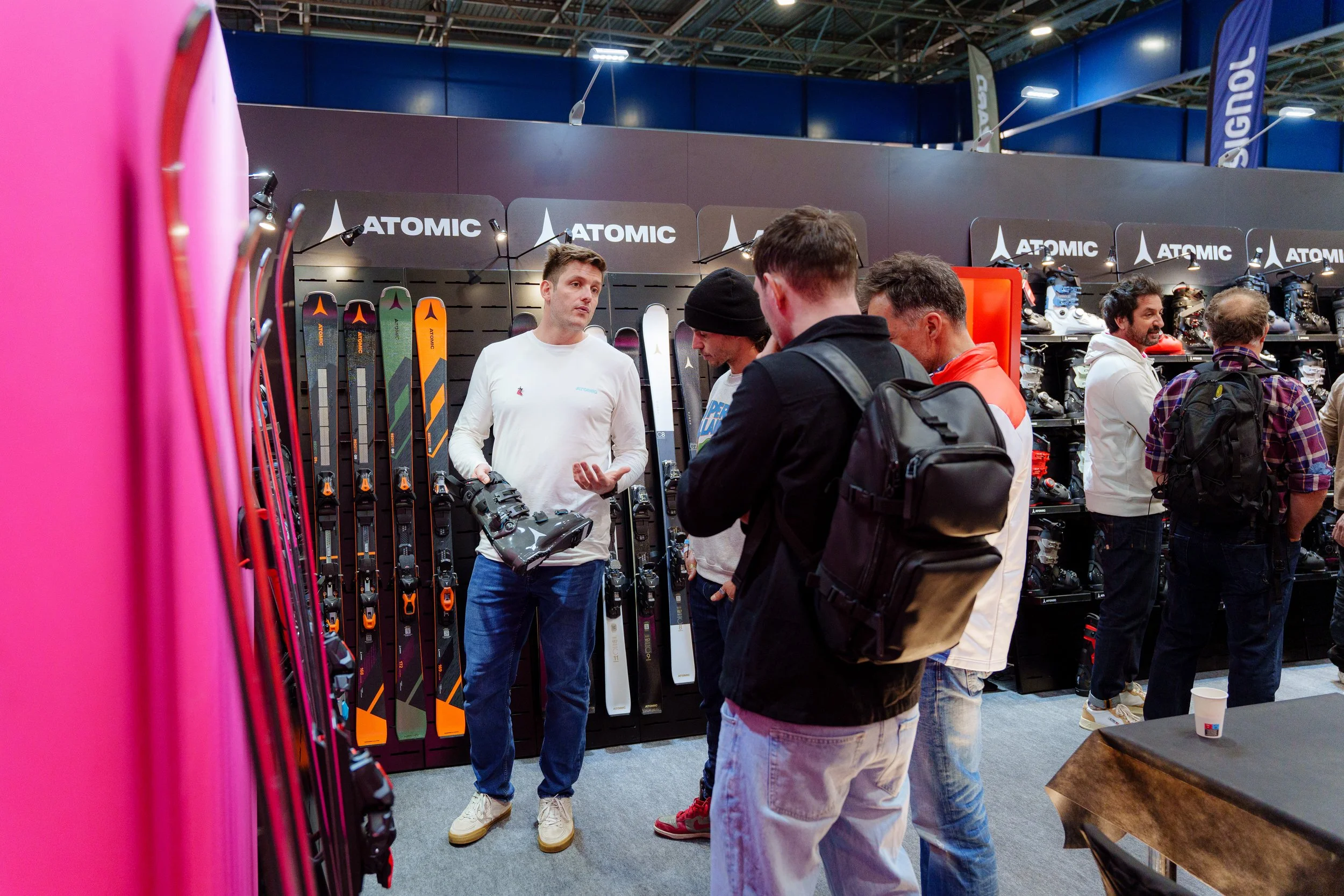 Un stand de présentation de skis et fixations dans un salon, avec plusieurs personnes discutant et regardant les équipements de ski exposés.