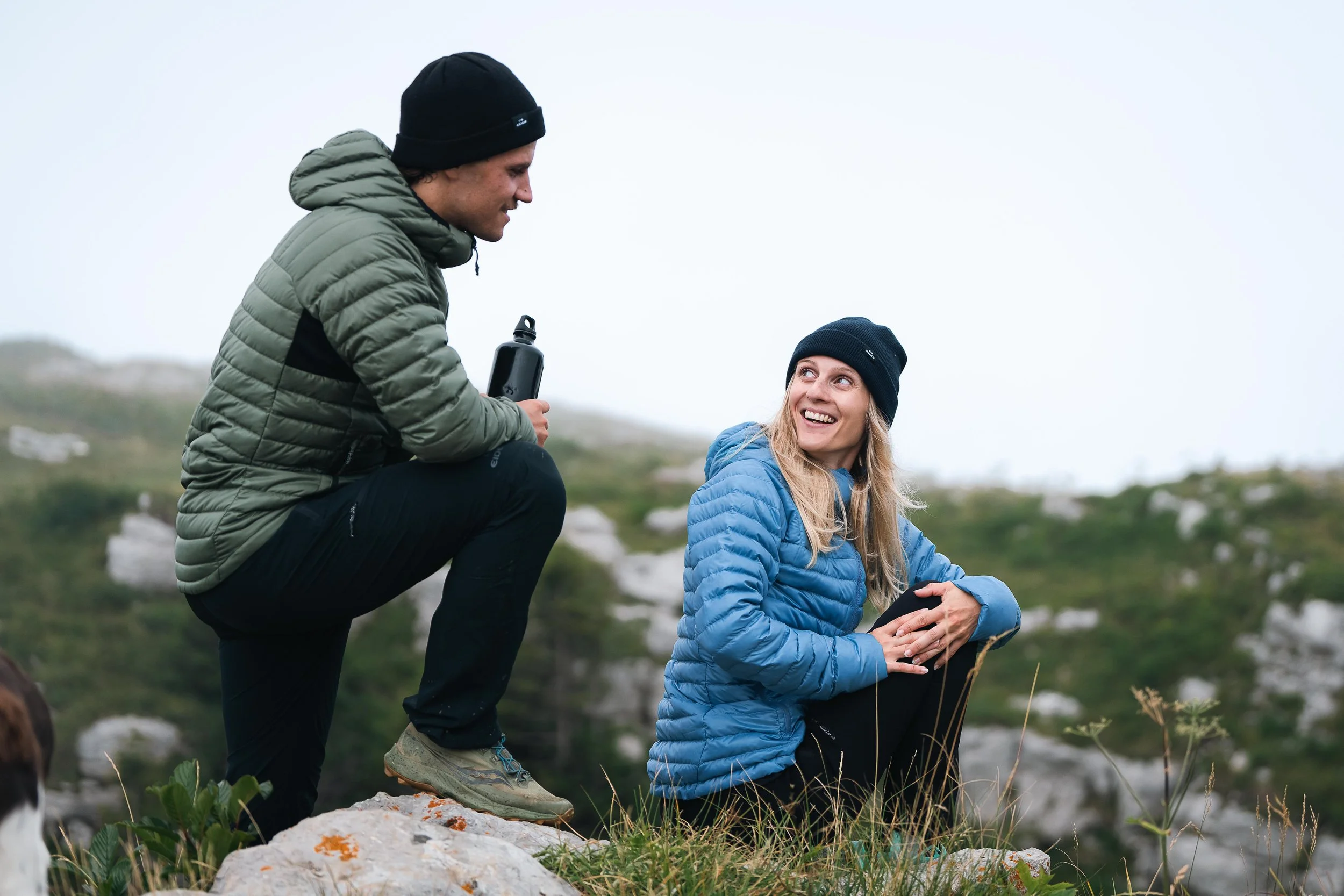 Deux personnes en tenues de randonnée discutant et souriant en plein air, l'une assise et l'autre accroupie tenant une bouteille d'eau, entourées de verdure.