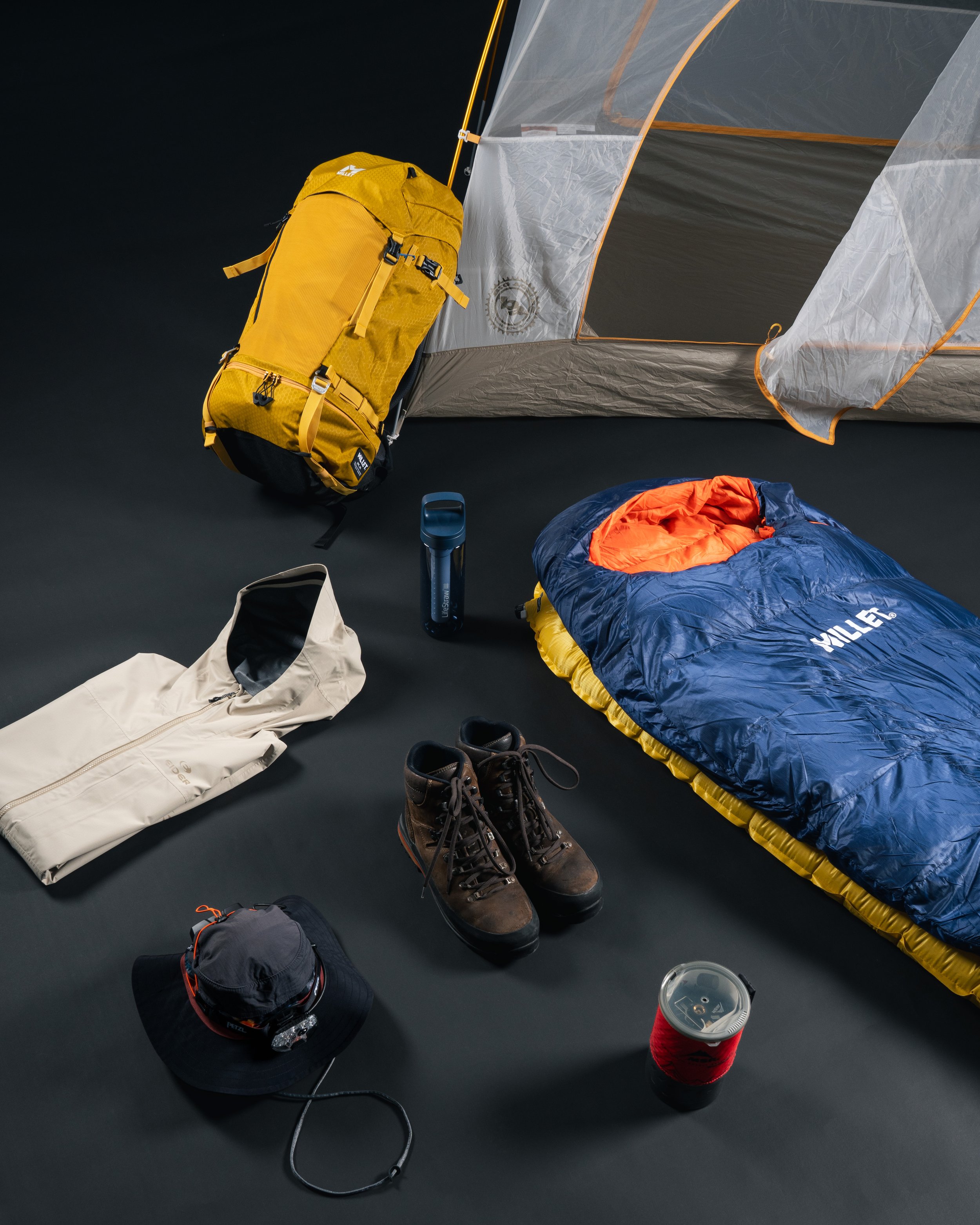 Équipements de camping, incluant un sac à dos jaune, une tente partiellement visible, un sac de couchage bleu et orange, une veste beige, des bottes de randonnée brunes, une tasse, une lampe frontale et un chapeau de randonnée.