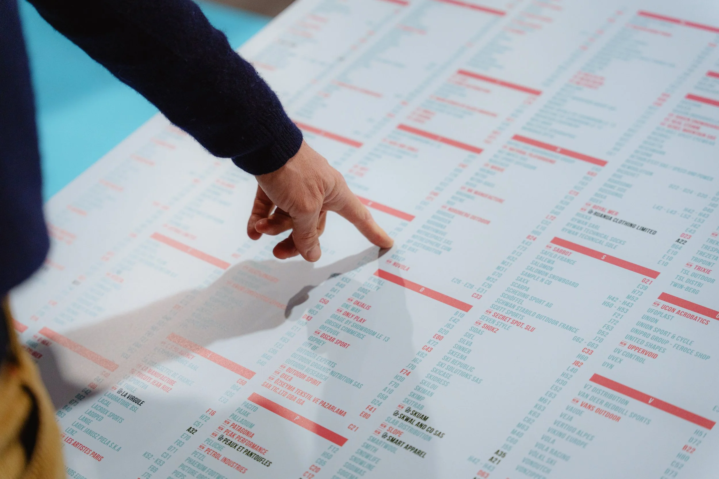 Une personne pointe du doigt un tableau avec des listes de noms et informations en rouge et bleu sur fond blanc.