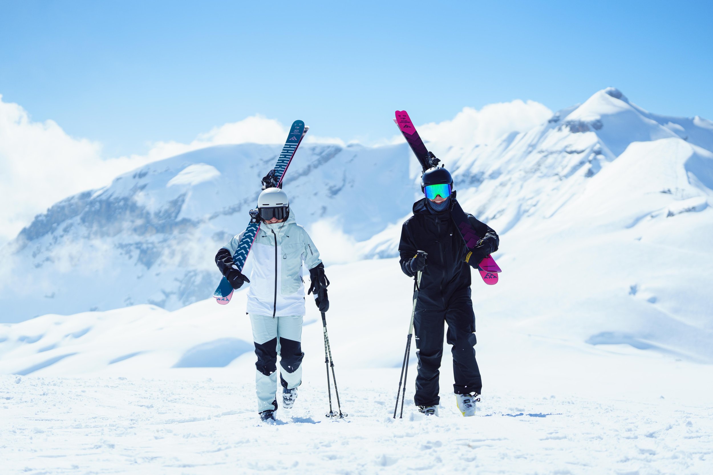 Deux personnes en tenue de ski marchent sur une piste enneigée avec des skis sur les épaules, montagne en arrière-plan.